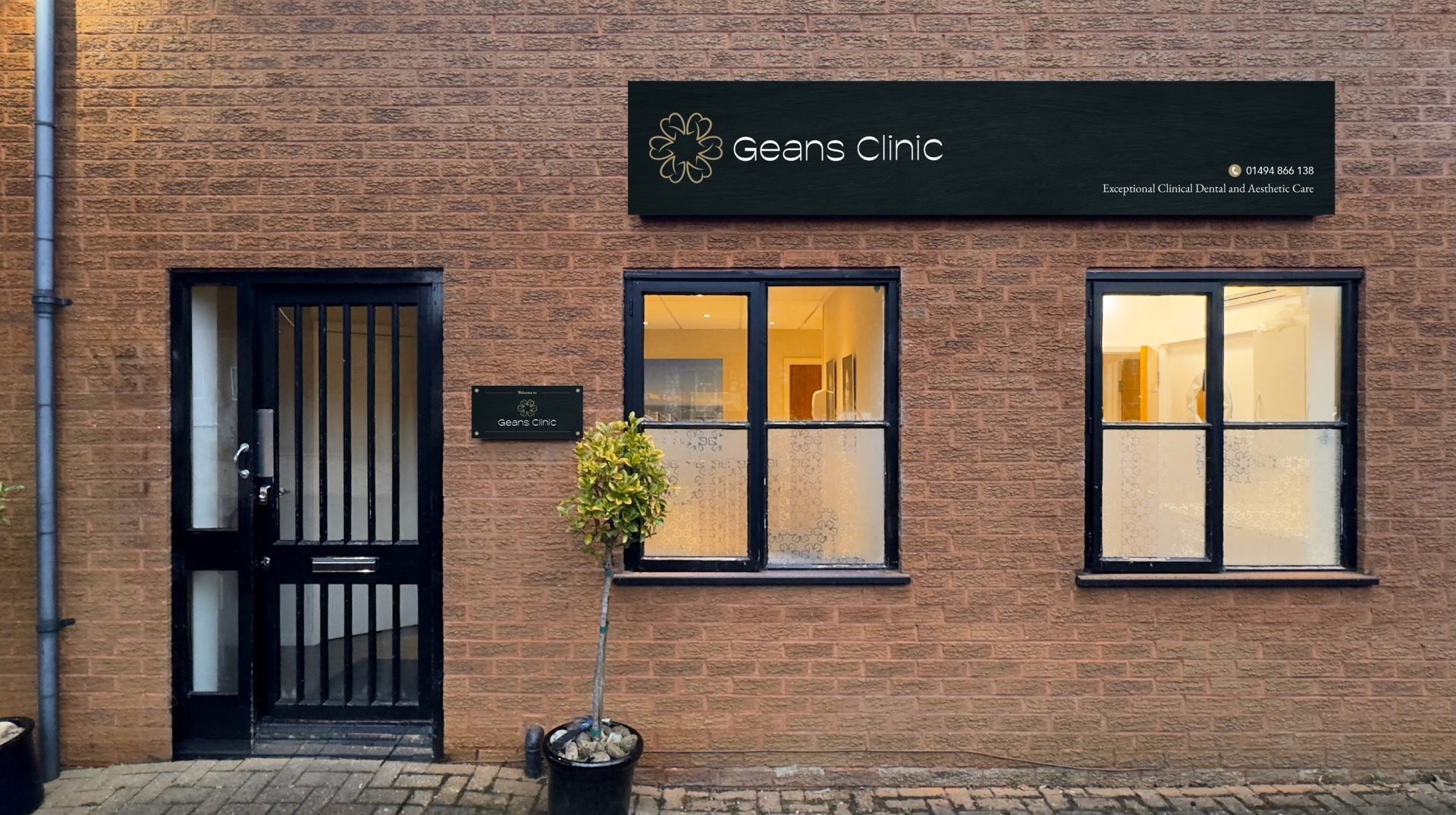 Brick building with a black door, two windows, and a sign that reads 'Geans Clinic'. There is a small potted tree outside and a sign on the wall next to the door. The clinic's sign features a flower logo and the words 'Geans Clinic' along with a phon