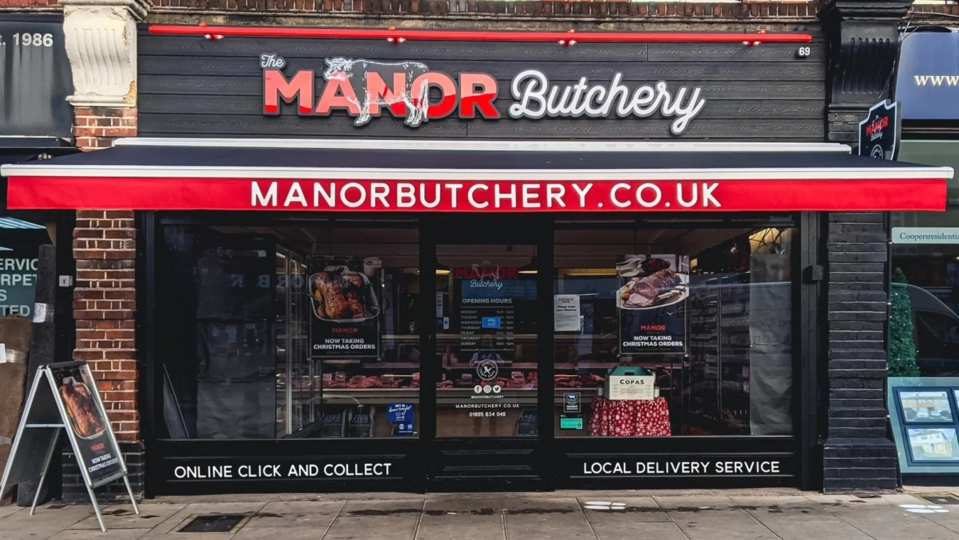 Front view of a butcher shop named 'The Manor Butchery', with signage displaying the website manorbutchery.co.uk. The shop has a black exterior with glass display windows showing meat products, and red and white signage indicating online ordering and