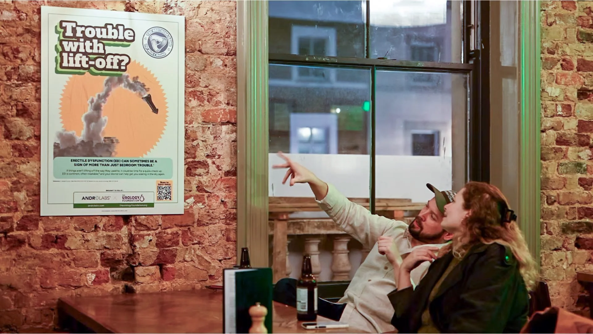 Two people sitting at a table in a brick-walled room, engaging in a conversation, with a poster about erectile dysfunction on the wall nearby.
