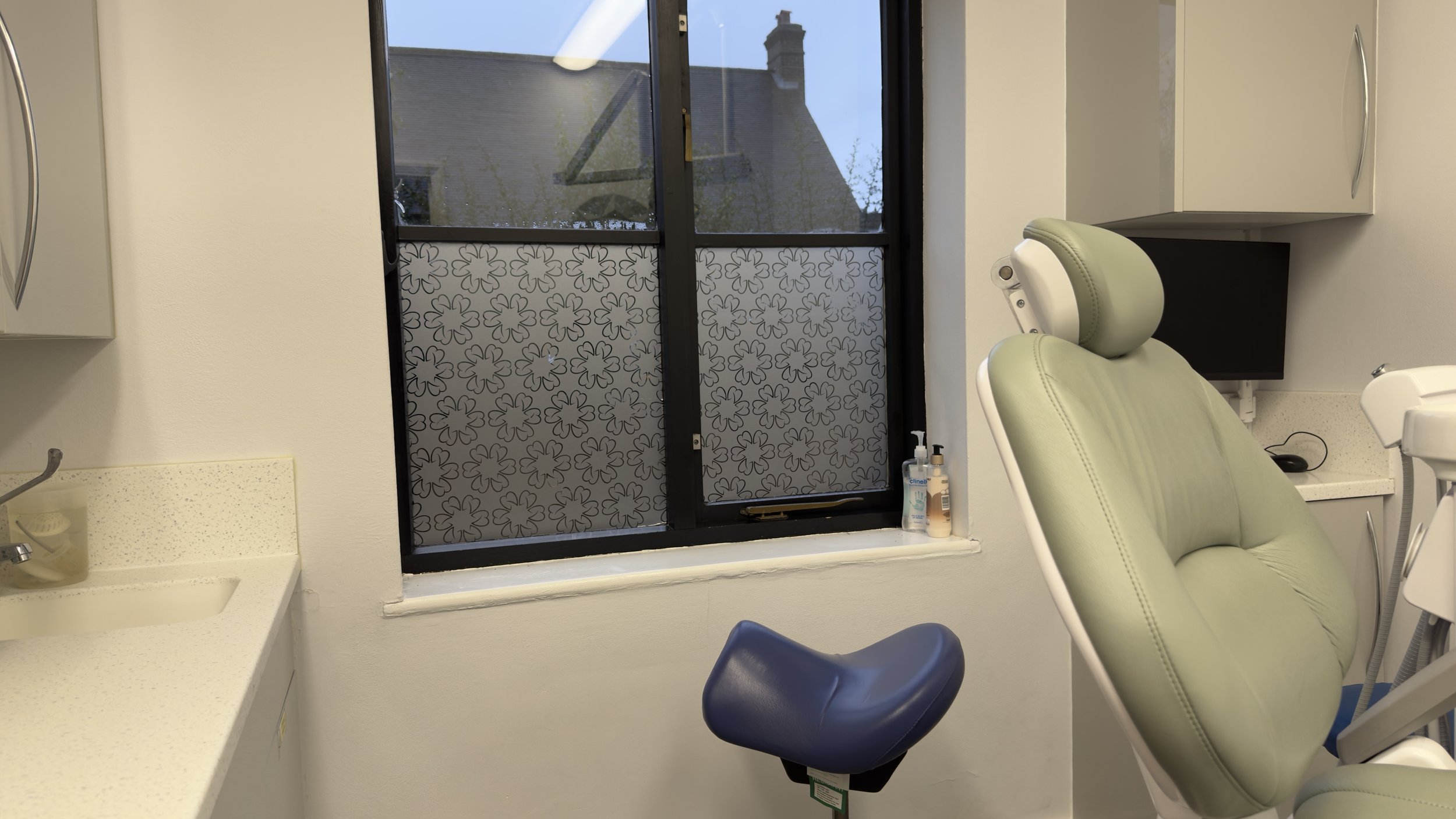 Medical examination room with a dental chair facing a window with patterned privacy film, a countertop with sanitizer and supplies, and cabinets.