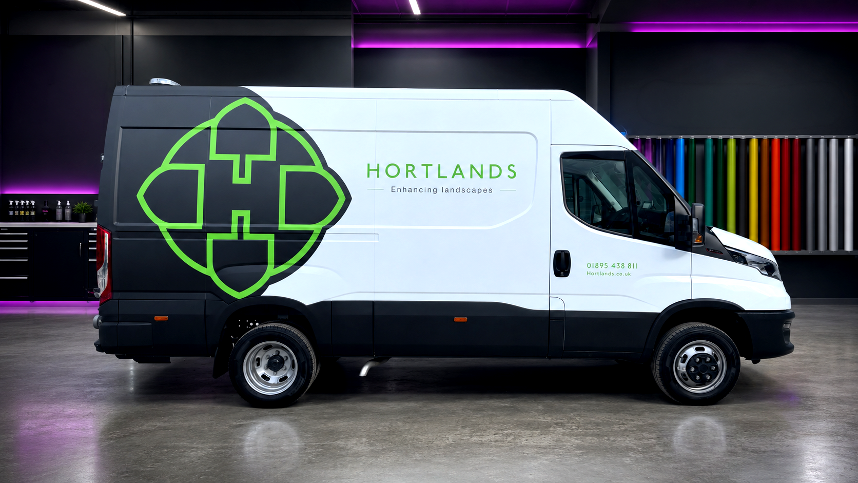 A white and black Hortlands landscaping delivery van parked indoors with a large green logo on the side and the text "HORTLANDS Enhancing landscapes". The background features a wall with colorful paint tubes and bottles on shelves.