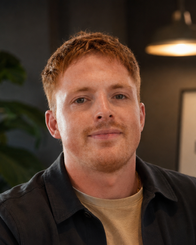 A young man with red hair and a slight smile, wearing a black jacket over a beige shirt, sitting indoors near a large green plant and a wall with a motivational poster.
