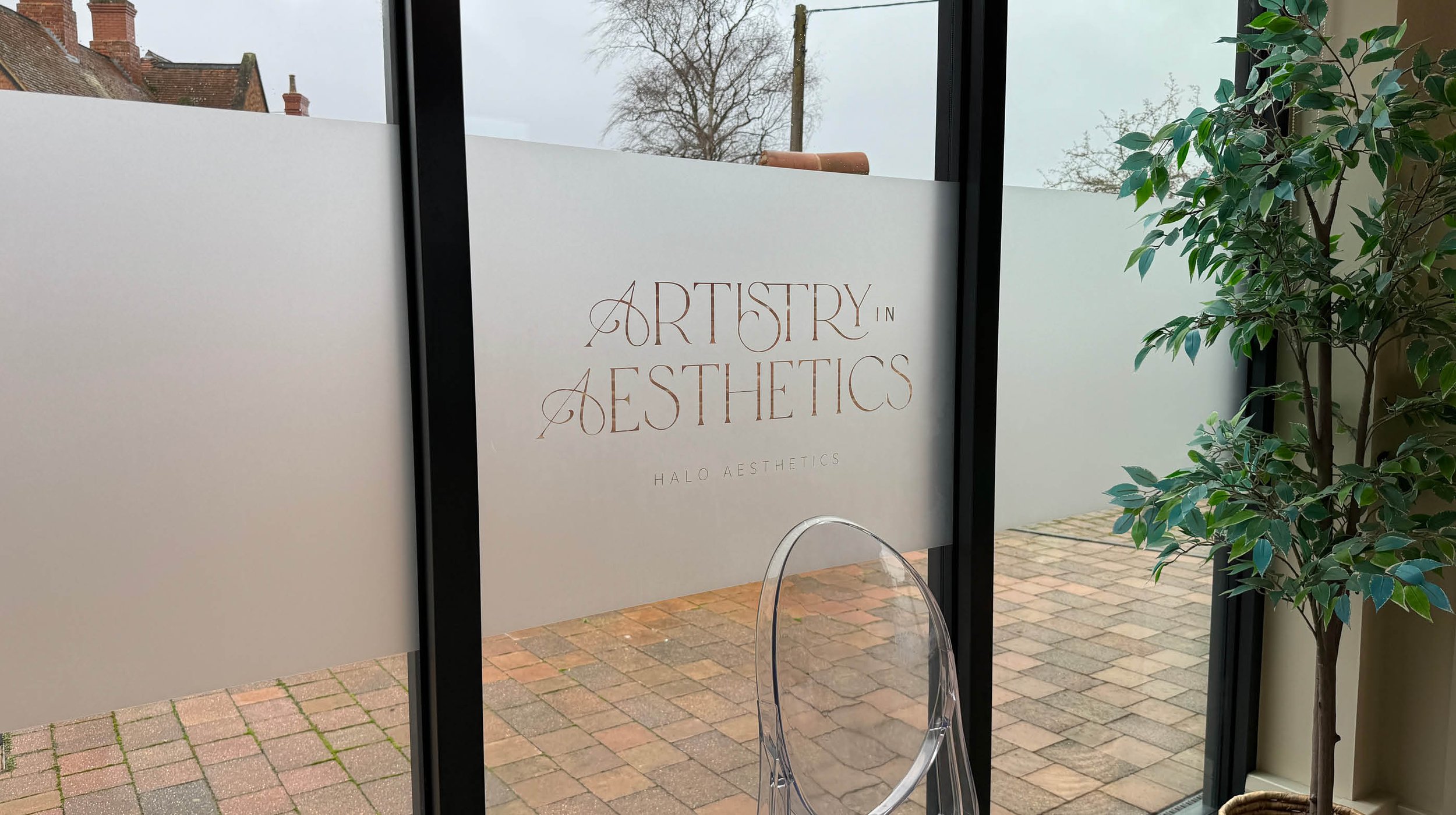 Sign on glass door reads 'Artistry in Aesthetics, Halo Aesthetics'. Outside view of brick pavement, a brick building with chimneys, and leafless trees.