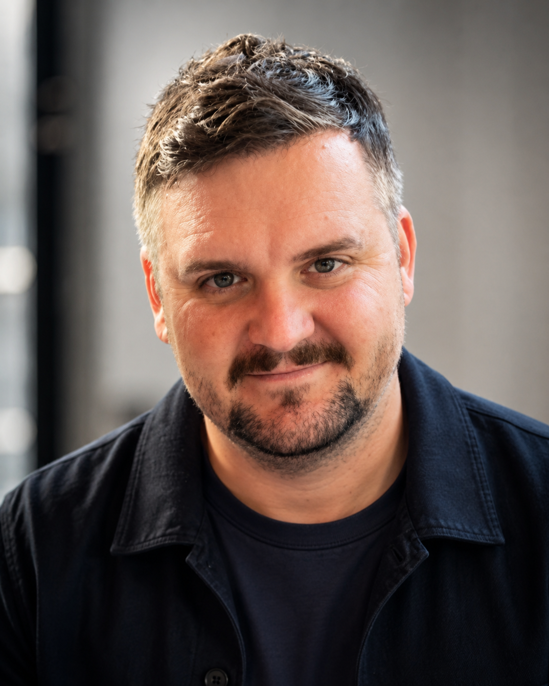 Close-up of a smiling man with short, grayish-brown hair, a short beard, and blue eyes, wearing a black shirt with a black jacket, indoors with a blurred background.