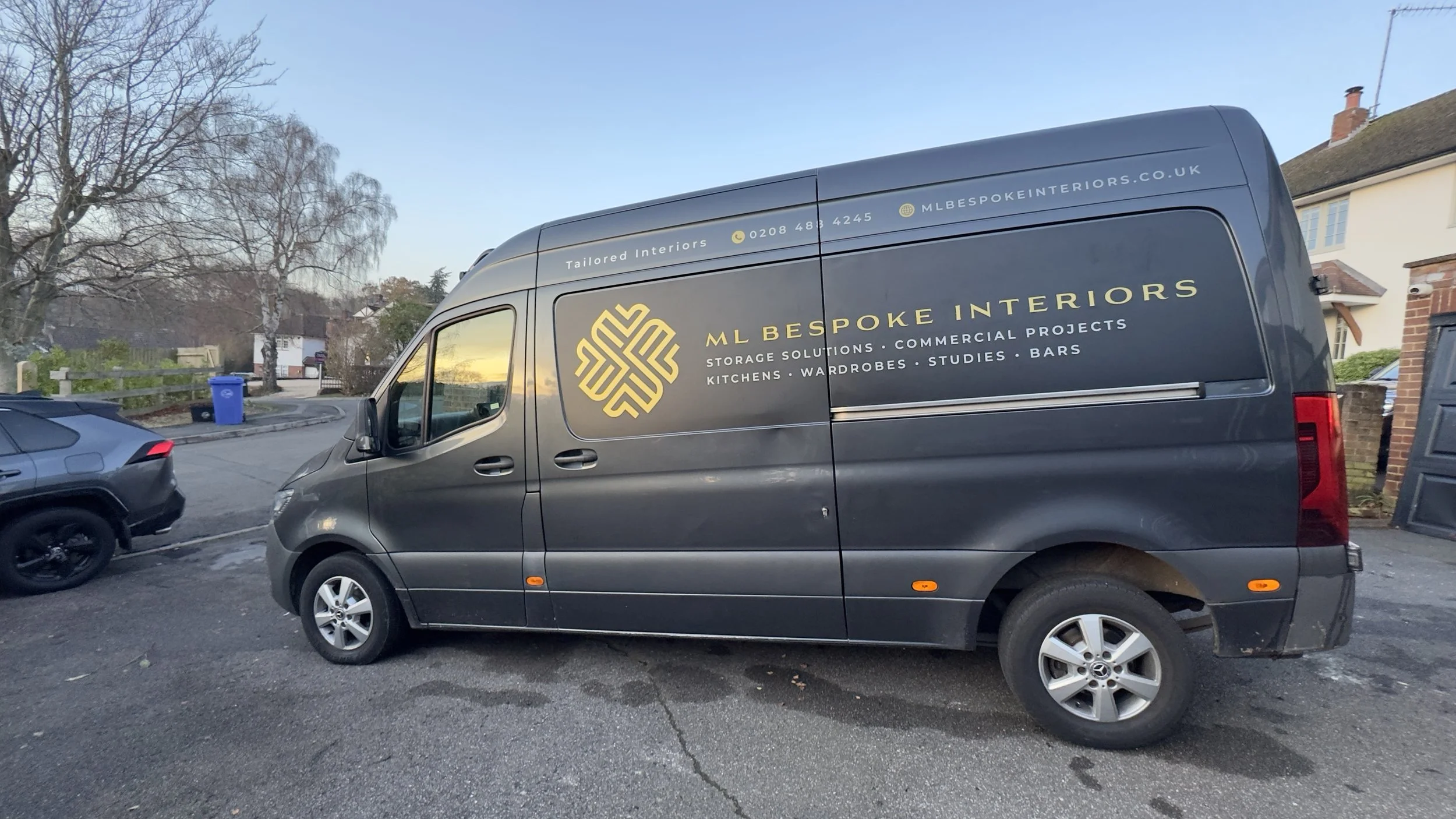 A dark gray van parked on a residential street, with two other cars nearby and trees in the background. The van displays the logo and contact information for ML Bespoke Interiors, a company specializing in storage solutions, commercial projects, kitc