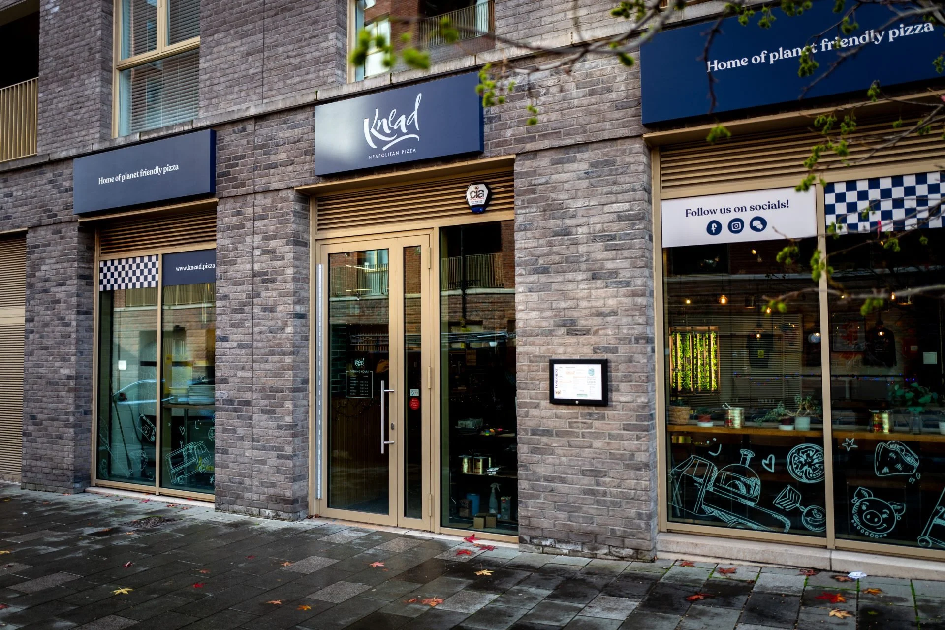 Exterior view of a brick restaurant with large windows and a blue sign that reads 'Knad Neapolitan Pizza.' There is also a sign saying 'Home of planet friendly pizza' and social media icons on the window.