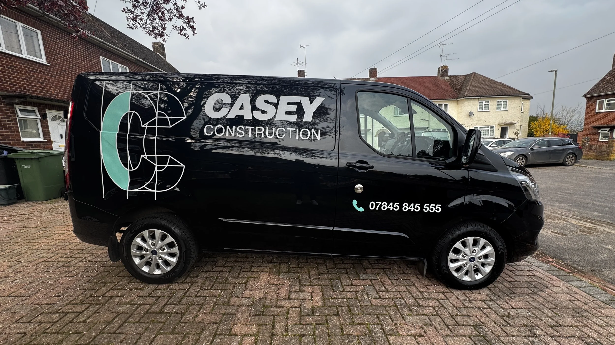 A black van with the company name 'CASEY CONSTRUCTION' and a phone number, parked on a brick driveway in a residential neighborhood with houses and cars visible in the background.
