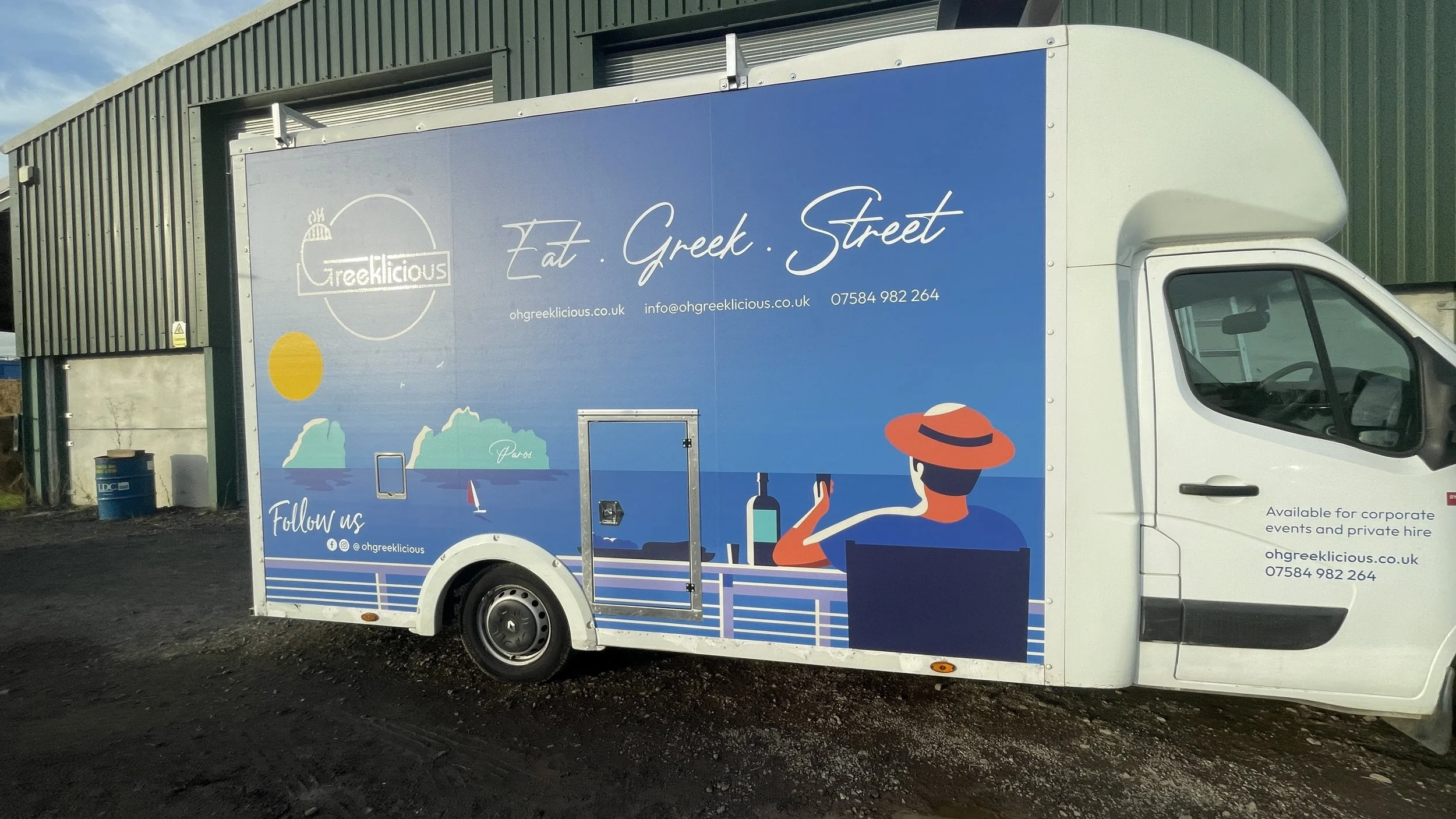 A food truck with a blue graphic wrap advertising Greek cuisine, featuring a woman relaxing with a drink, a sun, mountains, and a sailboat in the background. The text on the truck reads "Eat Greek Street" and includes contact information and social m