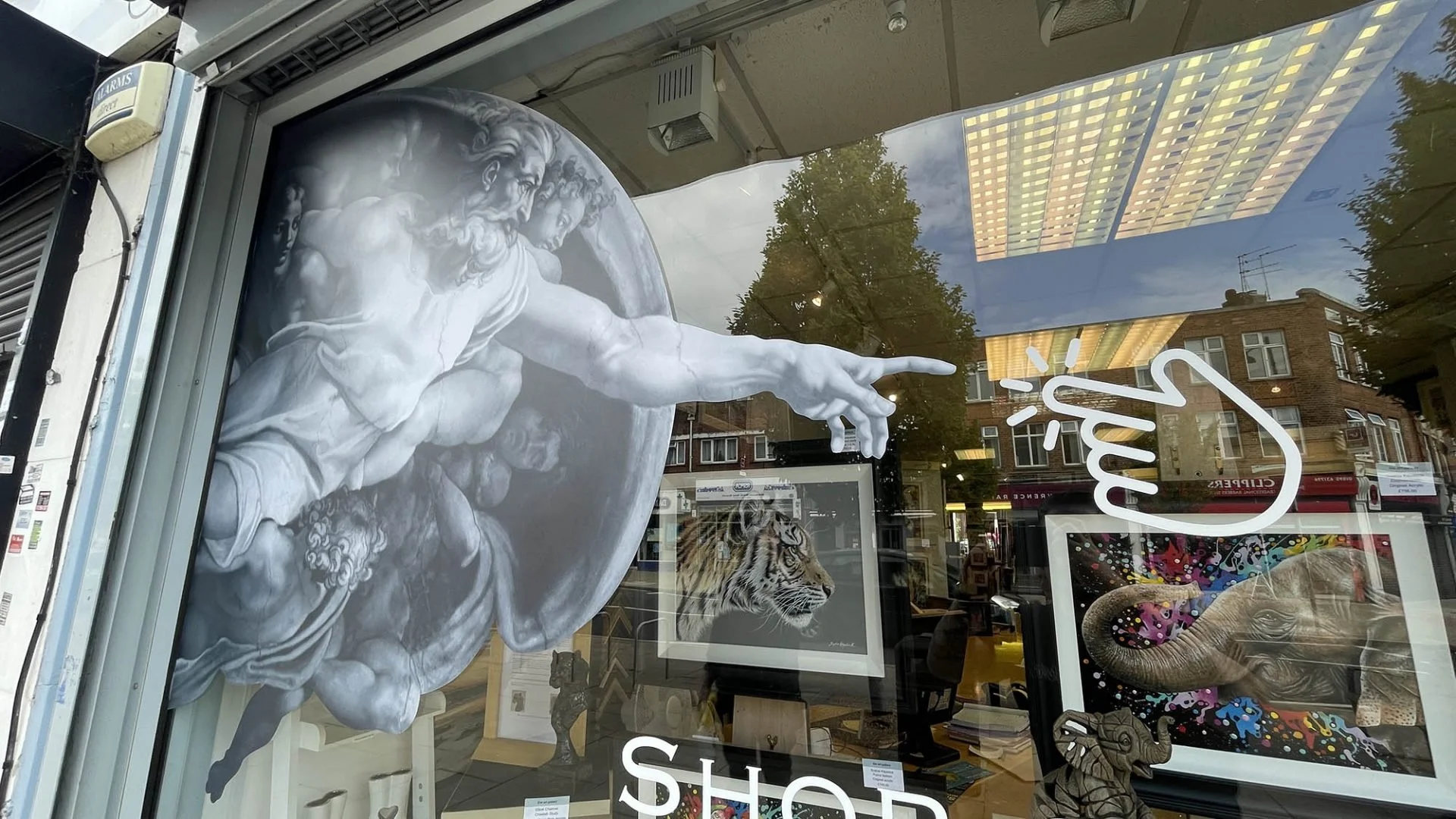 A storefront window display featuring a large white sculpture of Michelangelo's 'The Creation of Adam,' showing God reaching out to touch Adam's hand, with a tiger's photograph, colorful artwork of a snake, and a stylized thumb-up sign on the window.