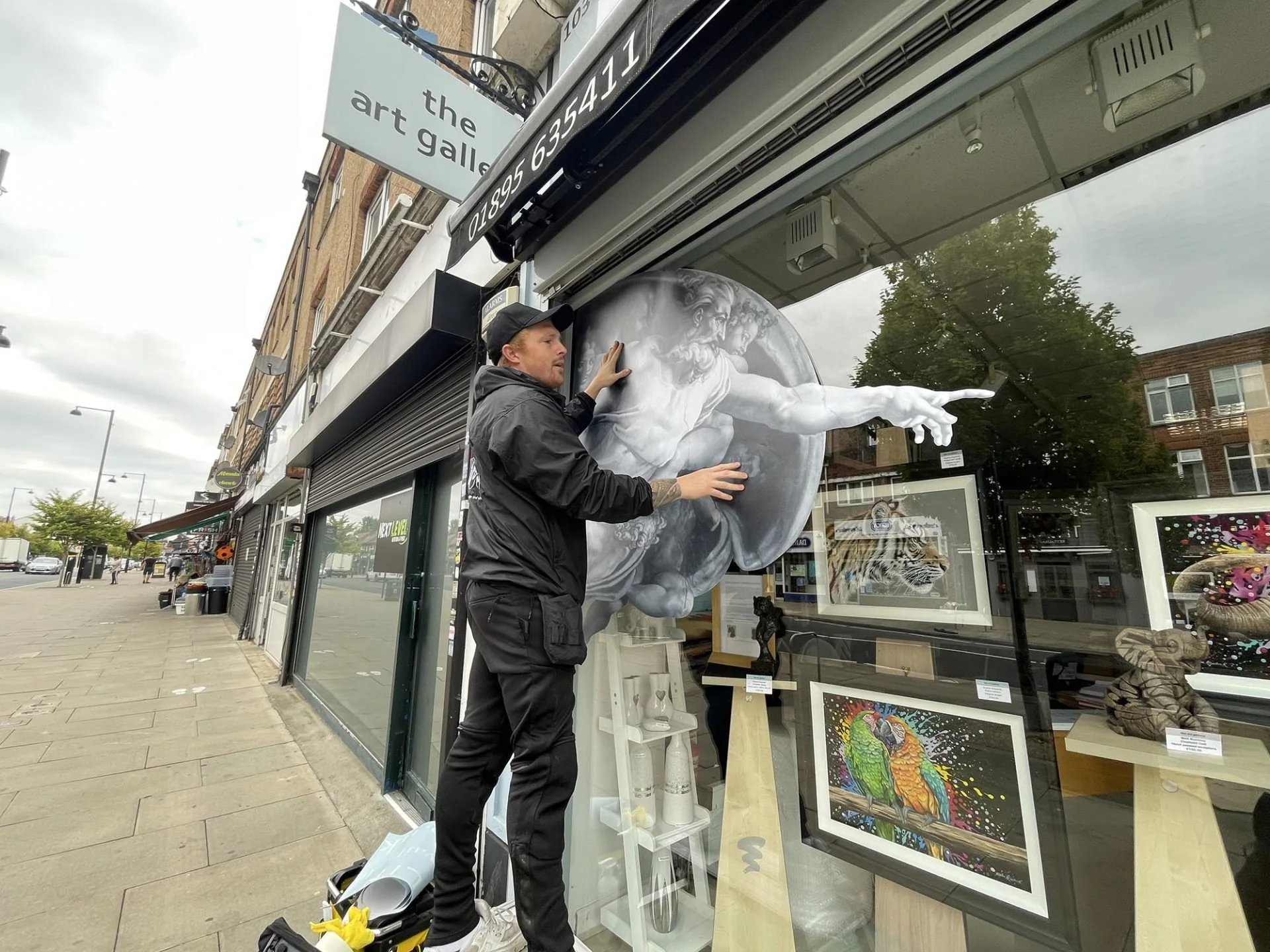 Man attaching a large black and white circular artwork of Michelangelo's 'The Creation of Adam' to the window of an art gallery storefront on a city sidewalk.