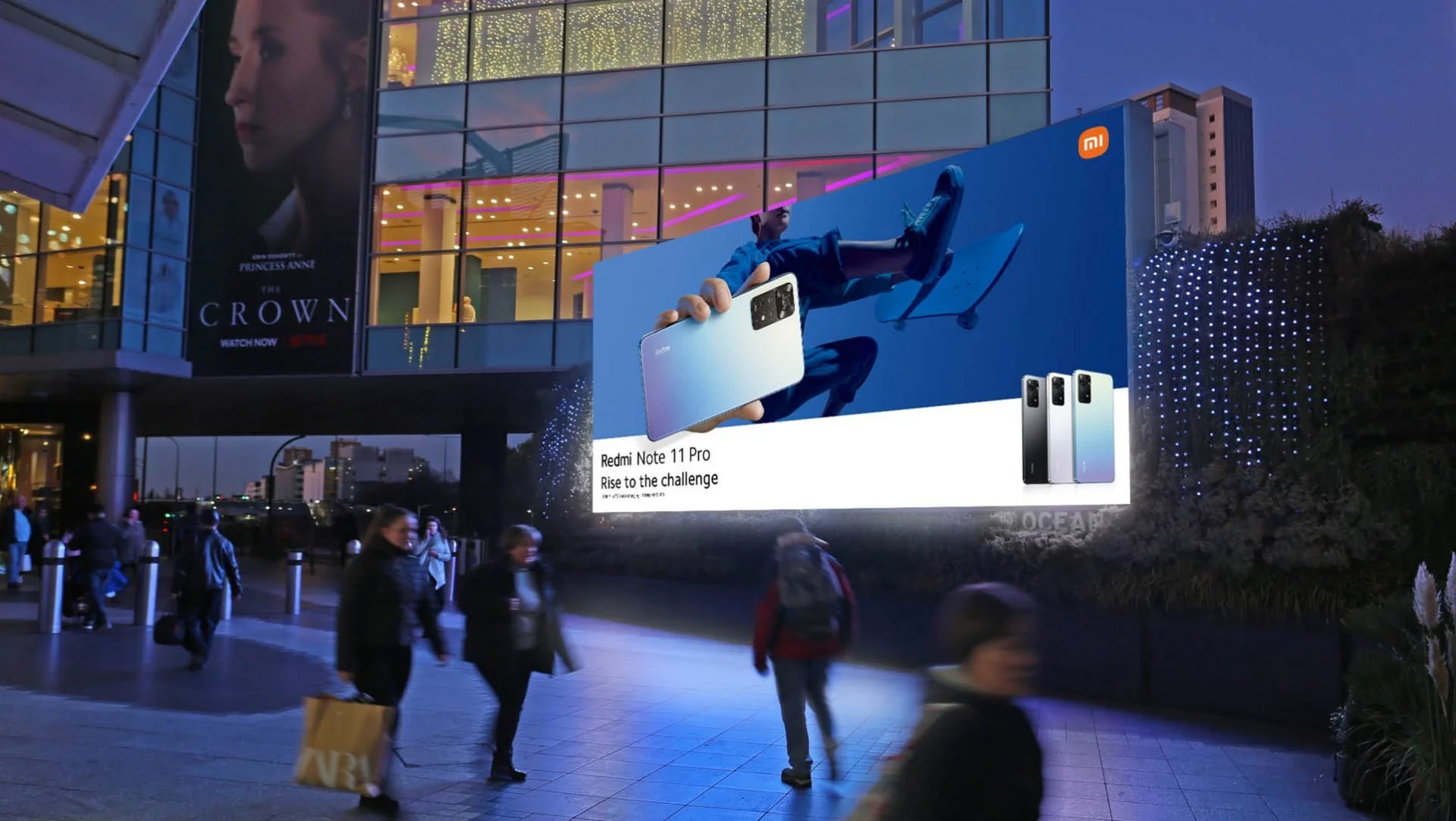 Large outdoor billboard advertising the Redmi Note 11 Pro smartphone with a tagline 'Rise to the challenge'. The billboard displays an image of the phone, and a man reclining review the phone. There are several shoppers walking past the billboard in a cityscape at dusk.