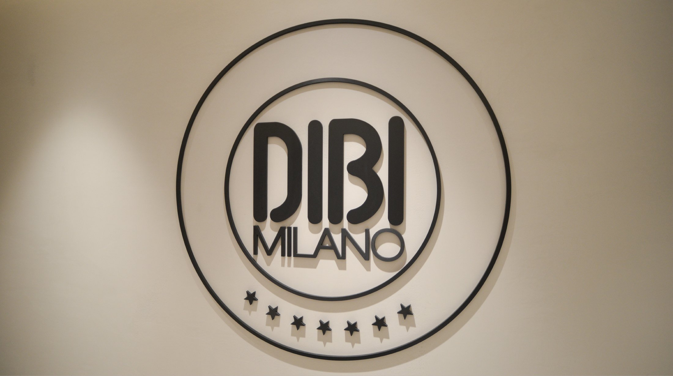 A circular sign that reads 'DUBI MILANO' with a decorative border and stars beneath it, mounted on a beige wall.