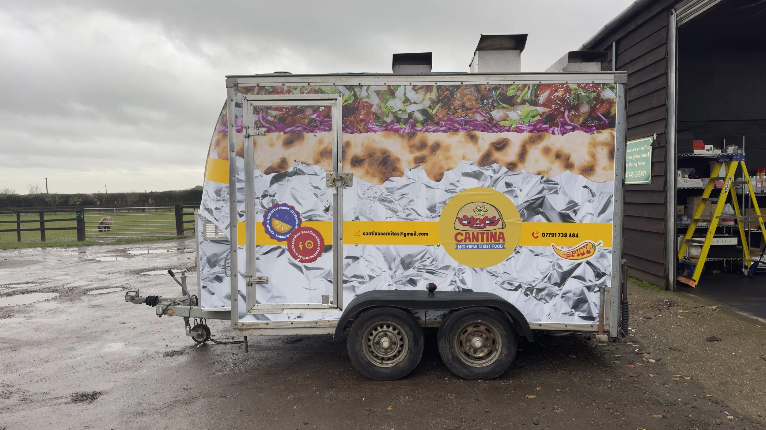 A food truck with colorful graphics of a taco and ingredients, labeled 'Cantina Mex-Fresh Street Food,' parked on a wet, muddy ground on a cloudy day, with a field and sheep in the background.