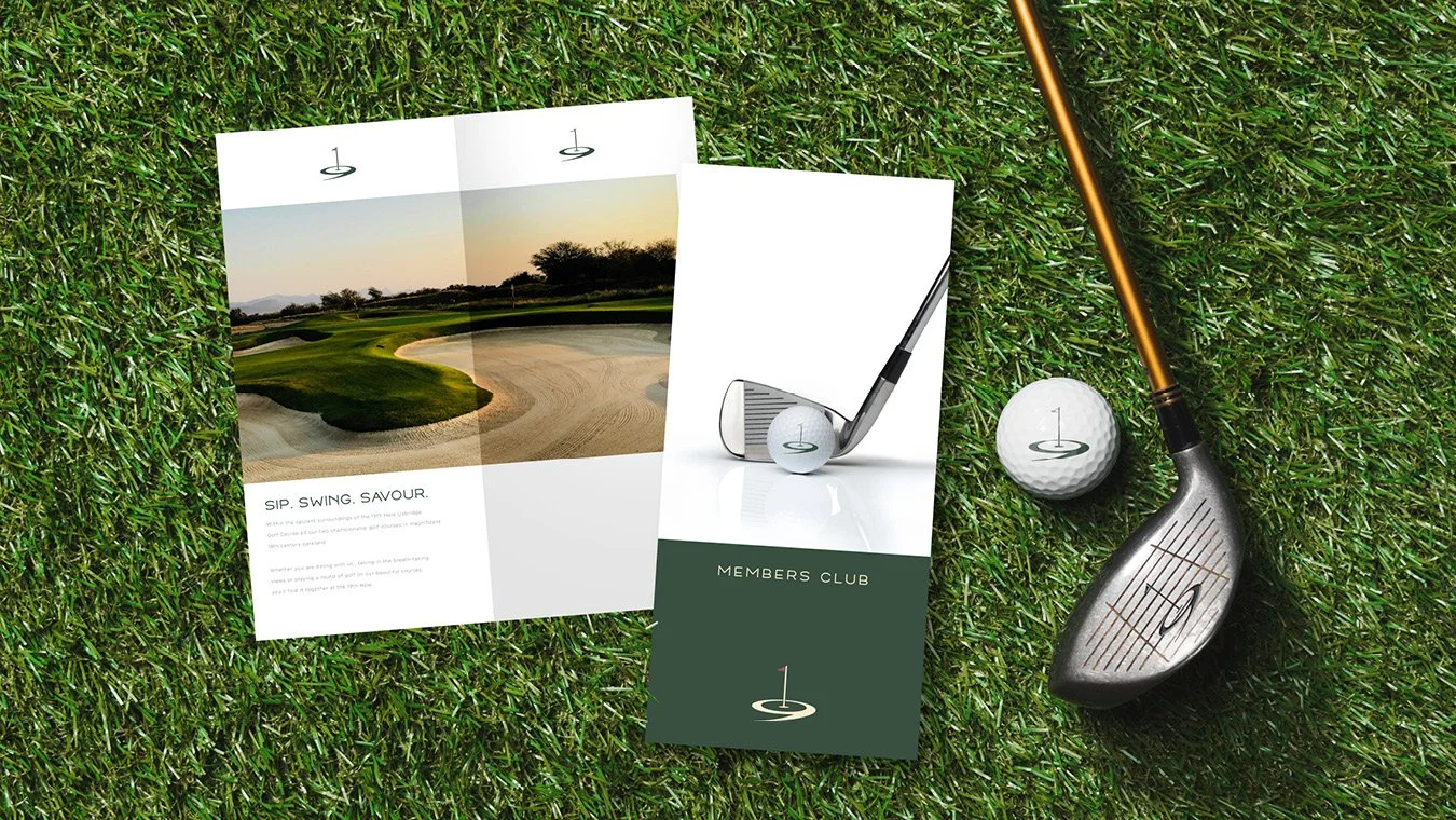 Golf equipment and promotional materials on grass, including a golf club, golf ball, golf tee, and brochures for a golf club, with scenic golf course photo.