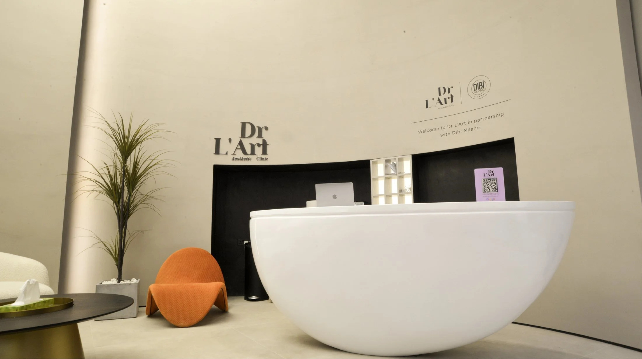 Reception desk at Dr L'Art aesthetic clinic with a modern white bathtub, a small orange chair, a green plant, and a black coffee table, with signage on the wall welcoming visitors in partnership with Dibi Milano.