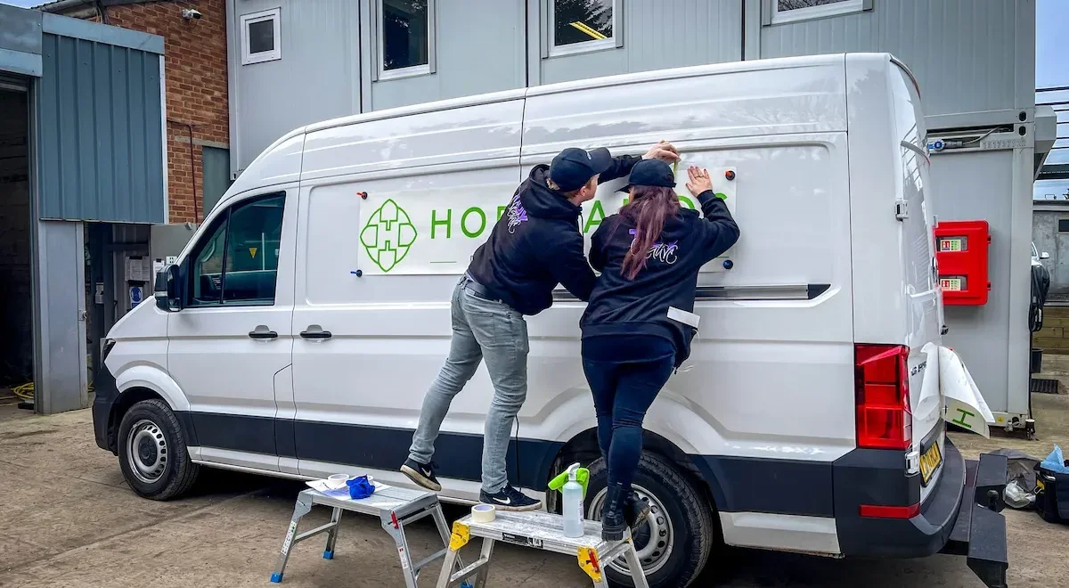 Two people installing a sign on the side of a white delivery van. They are standing on small ladders with tools and spray bottles nearby, working together to attach the sign.