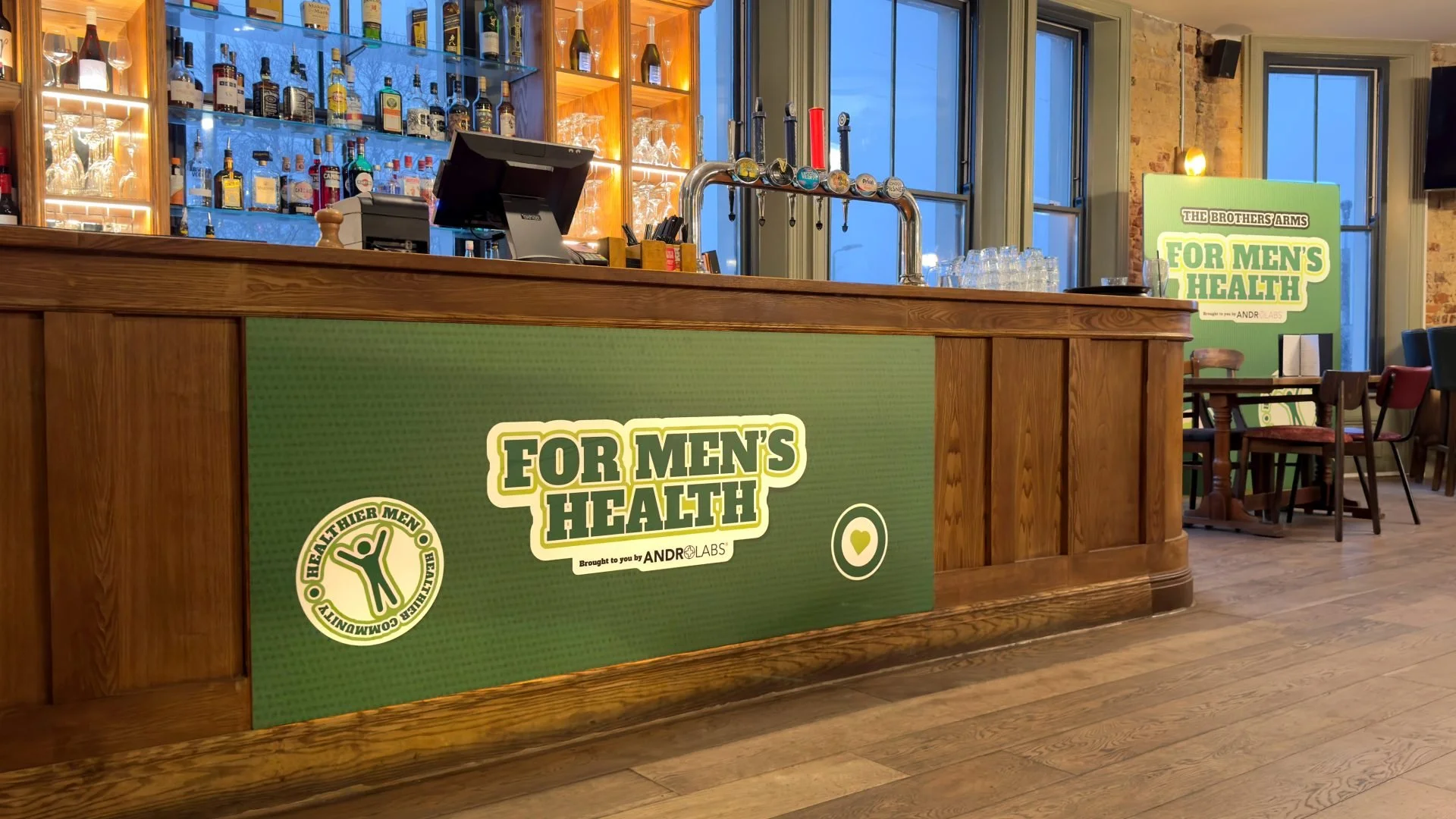 Interior of a bar or restaurant with a wooden counter and a green sign that reads 'For Men's Health,' emphasizing men's health awareness. Behind the counter is a well-stocked bar with various bottles and glasses, and large windows letting in natural 