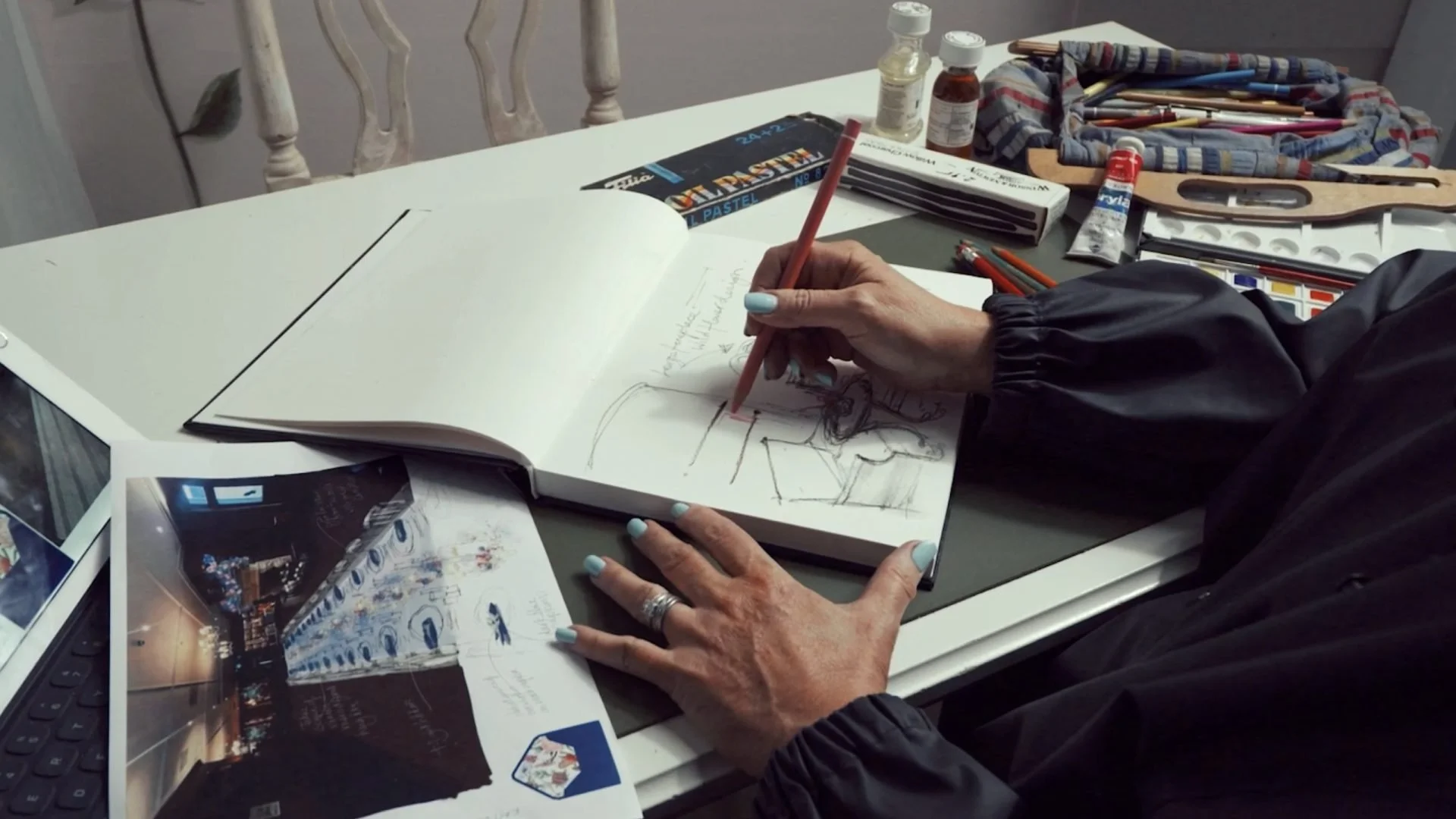 Person sketching in a notebook at a cluttered table with art supplies, brochures, and photos.