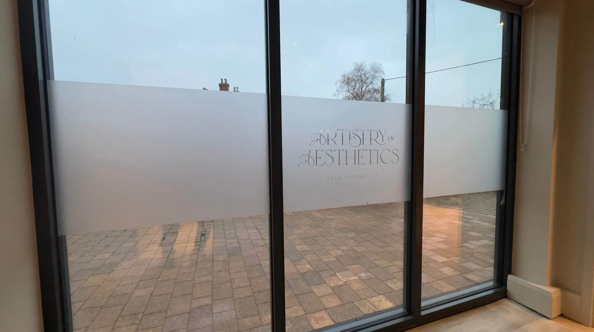 A sliding glass door with frosted glass covering about half of the window, showing an outdoor brick patio and a cloudy sky. On the frosted glass, there is text that reads 'AORTSTRY IN AESTHETICS HALO AESTHETICS'.