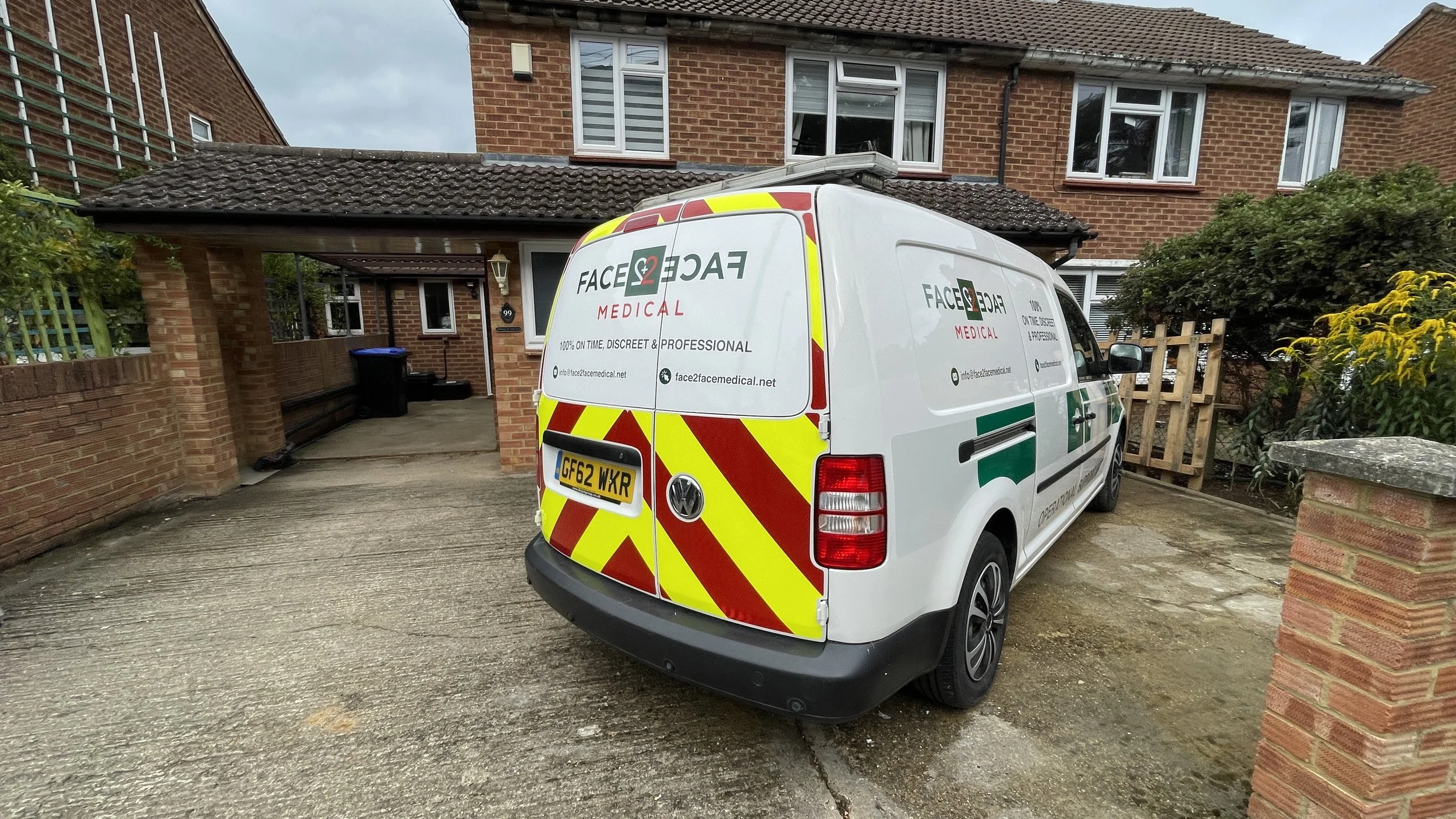 A white mobile medical van parked in a residential driveway in front of a brick house. The van has red and yellow reflective stripes and logos for Face2Face Medical, with contact information and website. The house has a covered porch, a blue recyclin