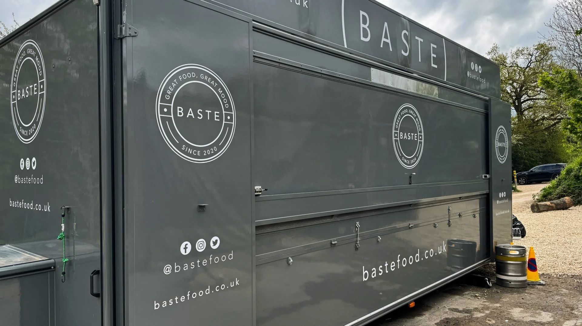 A large grey food truck with 'BASTE' logo and slogan 'Great Food, Great Mood' on the side. The truck has social media handles and website address 'bastefood.co.uk'. It is parked outdoors on a gravel area with trees and parked cars in the background, 