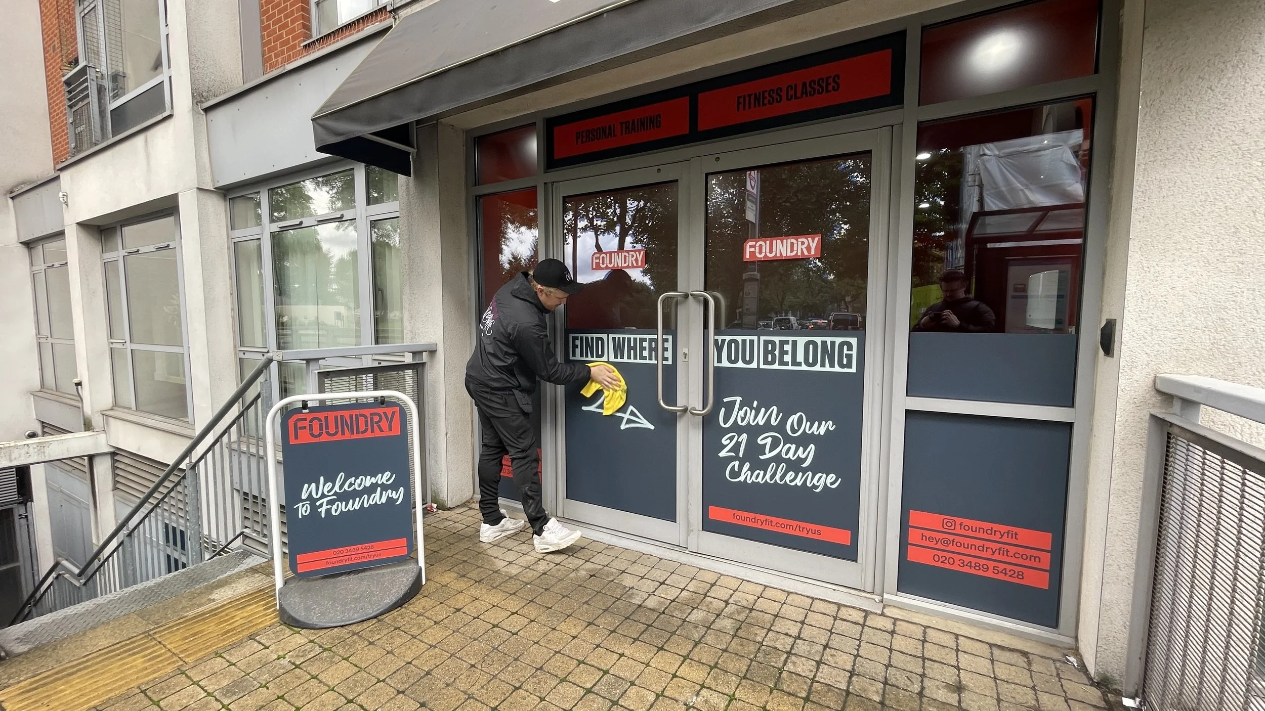 Man cleaning doors outside fitness gym called Foundry, which offers personal training and fitness classes. Sign mentions a 21-day challenge. The man is using a yellow cloth and wearing a black jacket and cap.