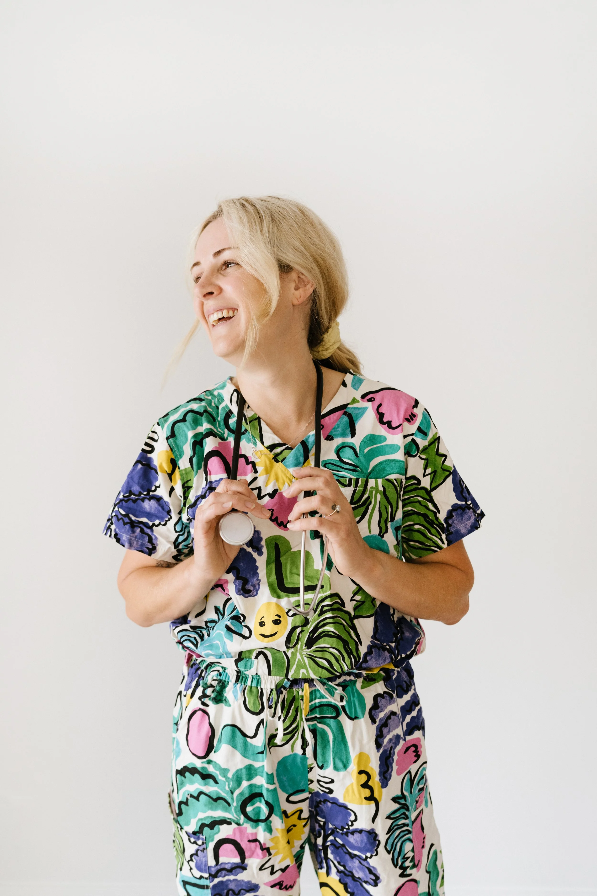 Beth Dewing RN with blonde hair is wearing colourful, patterned medical scrubs and holding a stethoscope, smiling and looking to the side against a plain white background.