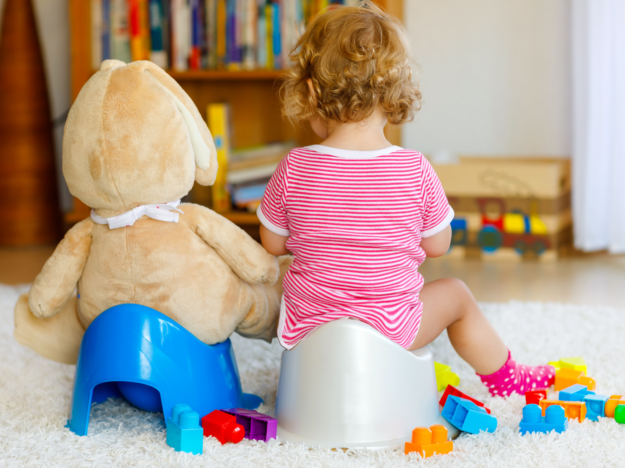 Child with curly hair sitting on a potty surrounded by colorful toys and a teddy bear in a playroom.