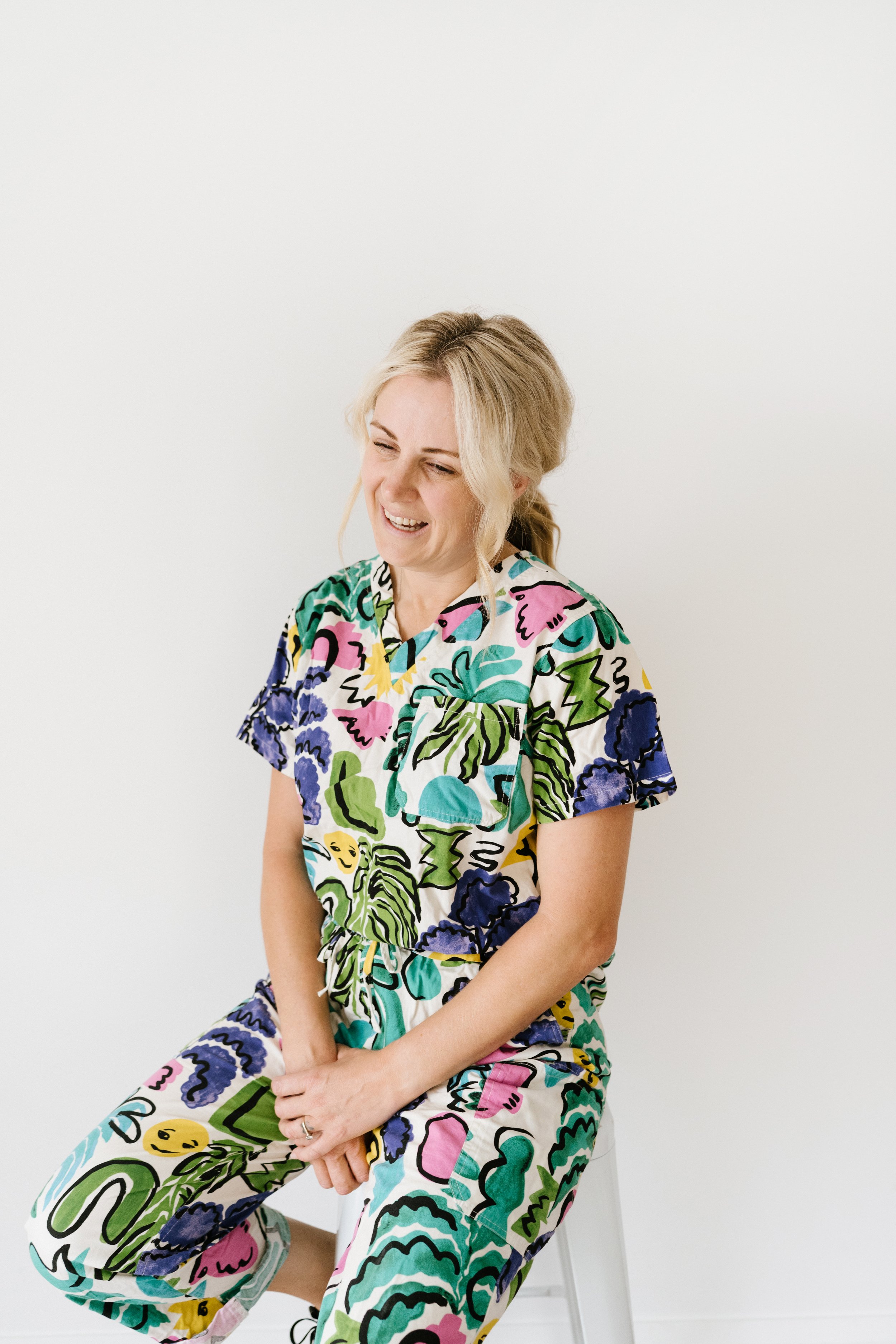 Beth Dewing RN with blonde hair smiling and looking down while sitting on a white chair against a plain white background. She is wearing a colourful, abstract-patterned outfit with bright colours and playful designs.