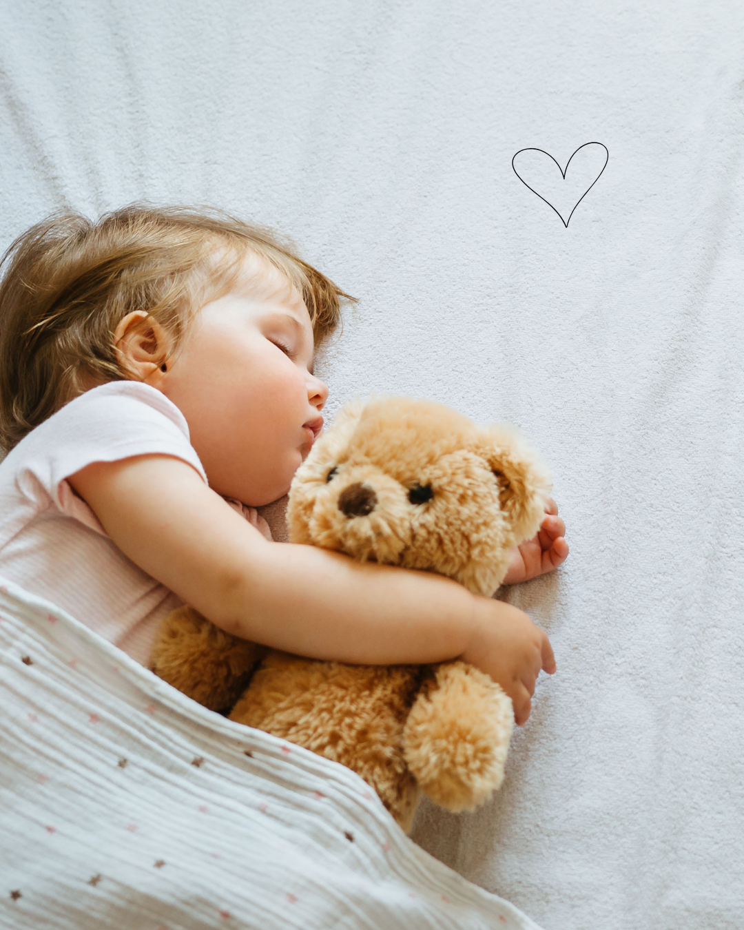 A young girl peacefully sleeping on her side, hugging a brown teddy bear, lying on a white bedding with a small drawn heart on the wall above.