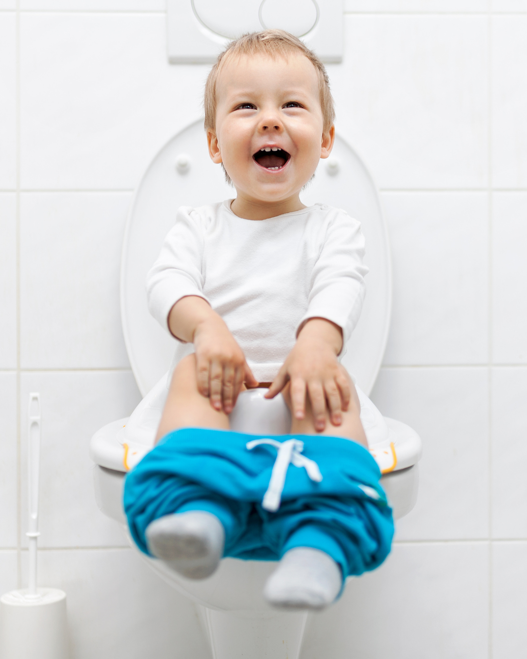 A young boy laughing and sitting on a toilet, wearing blue shorts and gray socks, in a bathroom with white tiled walls.