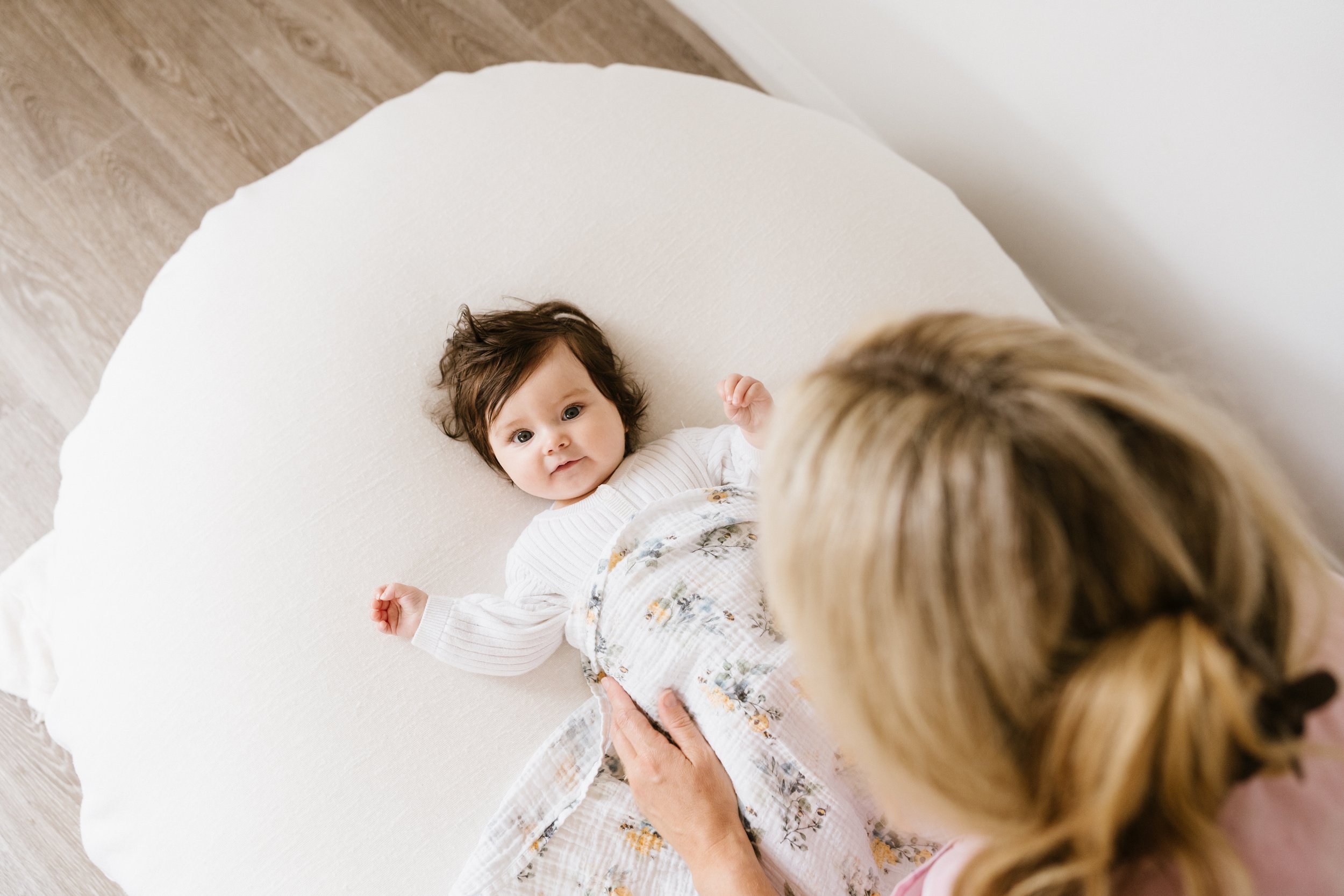 A baby with brown hair lying on a white blanket on the floor, looking up at Beth Dewing RN who is gently tucking the baby in.