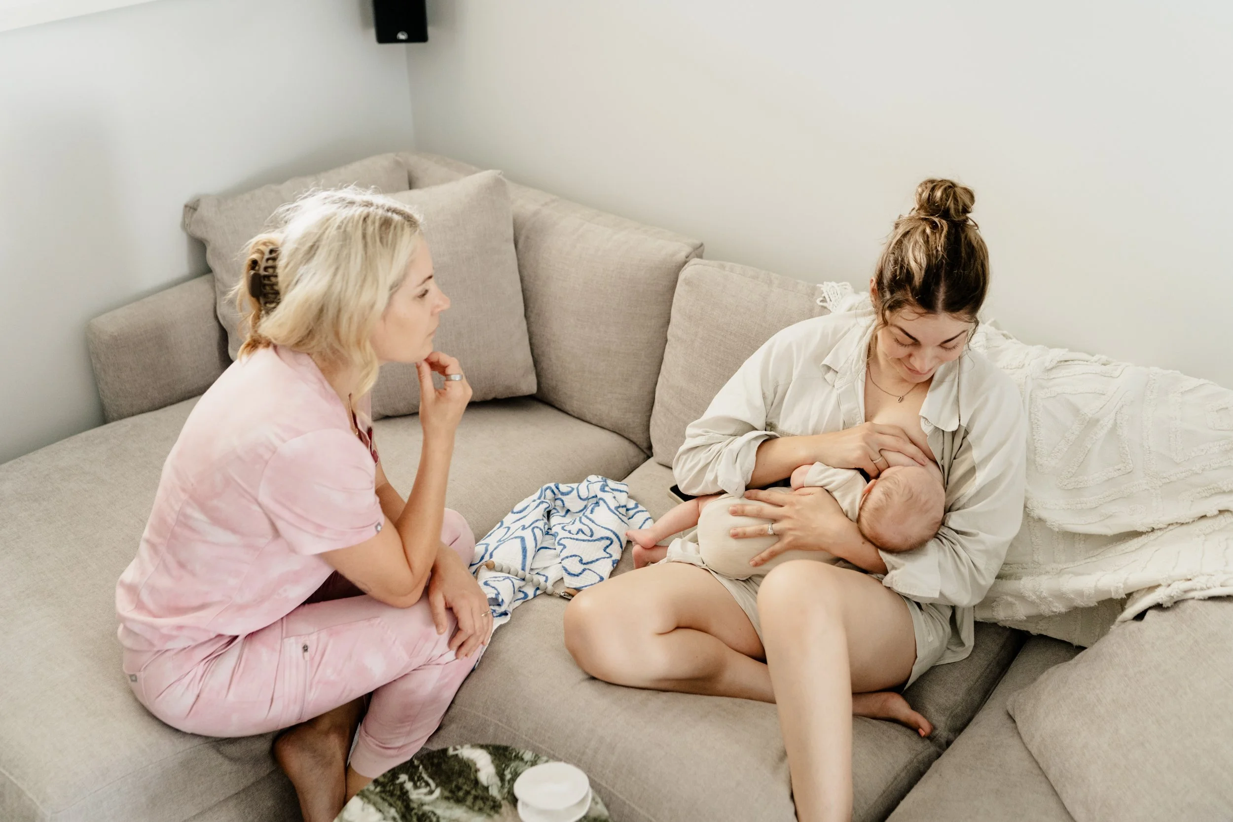 A woman in a white shirt is breastfeeding a baby while sitting on a couch, with Beth Dewing RN in pink scrubs sitting nearby completing a lactation consultation.