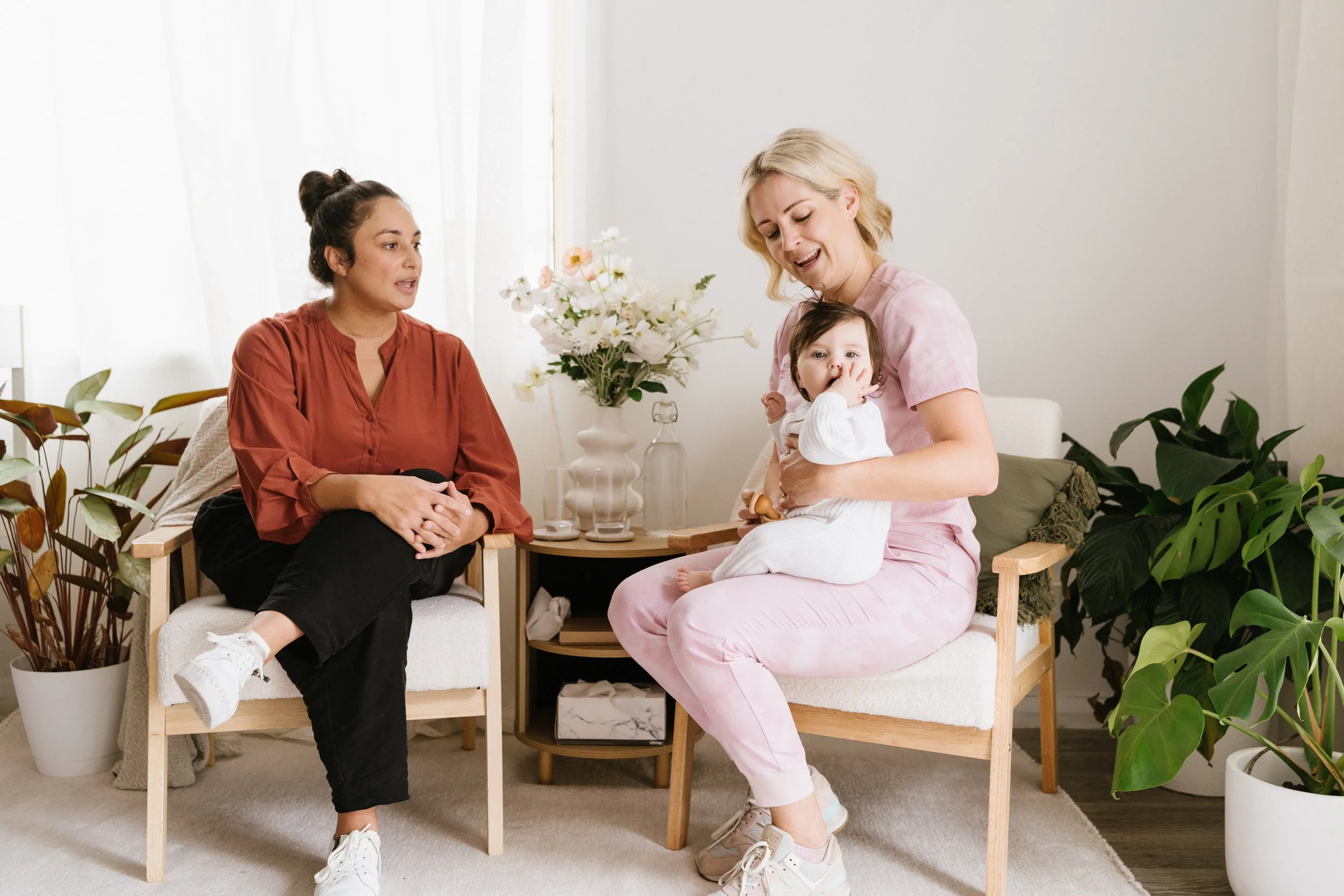 Beth Dewing RN with blonde hair in pink scrubs holds a baby with dark hair and a hand in their mouth, sitting on a wooden chair in a bright, plant-filled room, while a mum with dark hair in a bun and an orange top sits nearby listening.