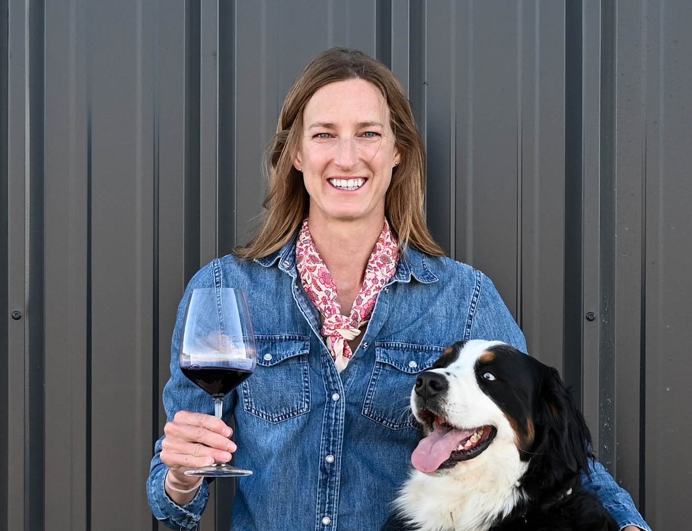 A woman with shoulder-length light brown hair, wearing a denim jacket and pink patterned scarf, smiling and holding a glass of red wine, sitting next to a black, white, and brown Australian Shepherd dog with its tongue out, against a dark gray metal wall background.