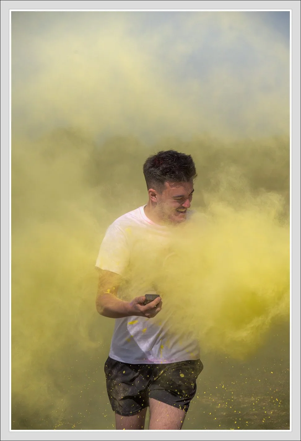 A young man in a white t-shirt and black shorts runs through a yellow powder explosion, smiling and holding a phone in his right hand.