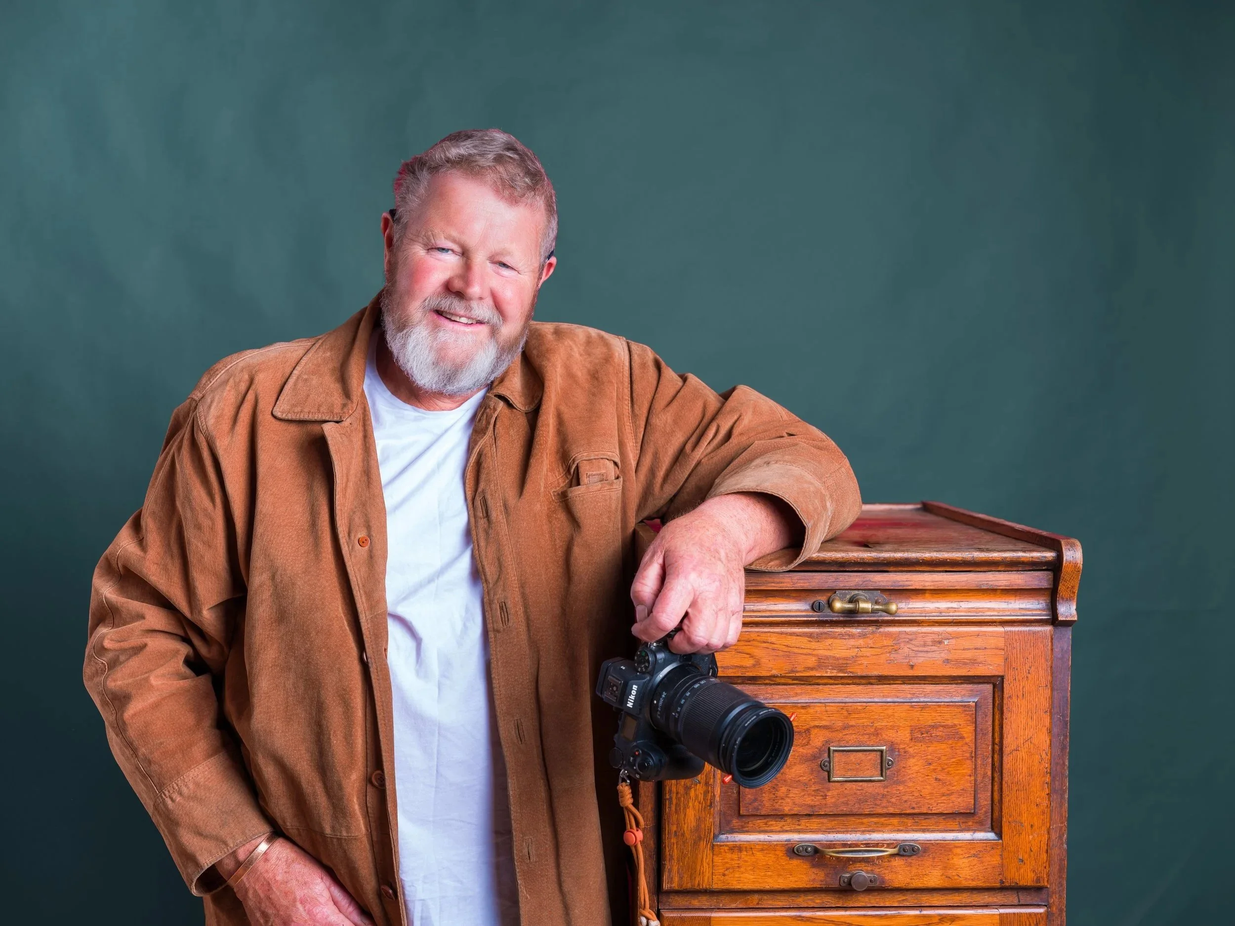 An older man with a white beard smiling, wearing a brown jacket and white shirt, holding a camera, standing next to a wooden drawer chest against a green background.