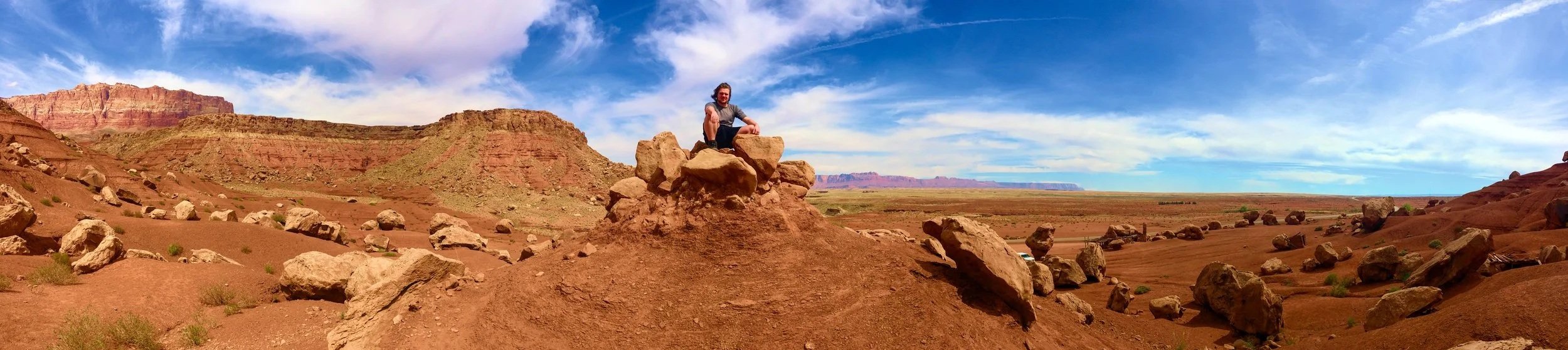 A person sitting on a large rock formation in a desert landscape with red rock formations and a blue sky with clouds.