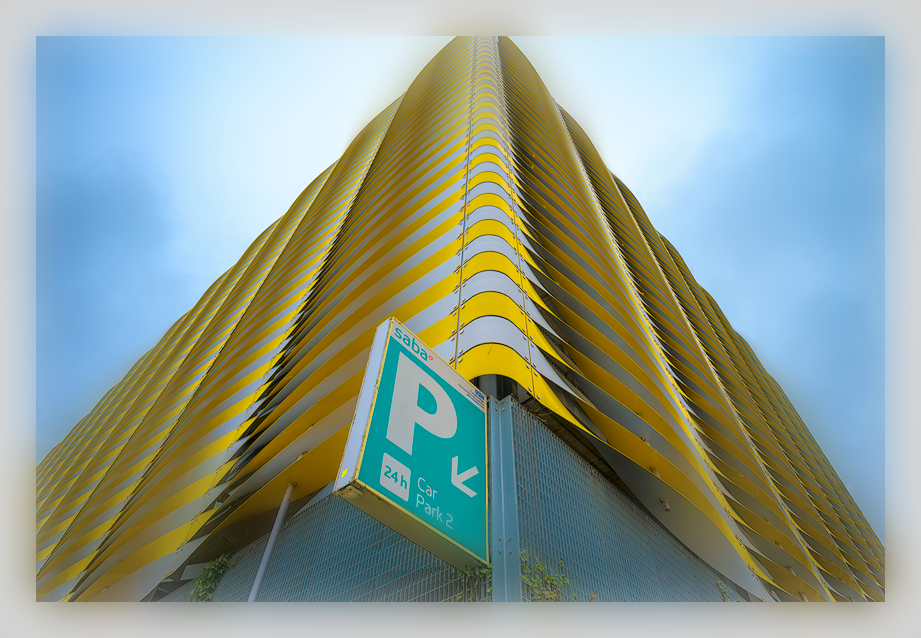 Looking up at a modern multi-story parking garage with yellow and black striped balconies, with a parking sign in the foreground indicating 24-hour parking.