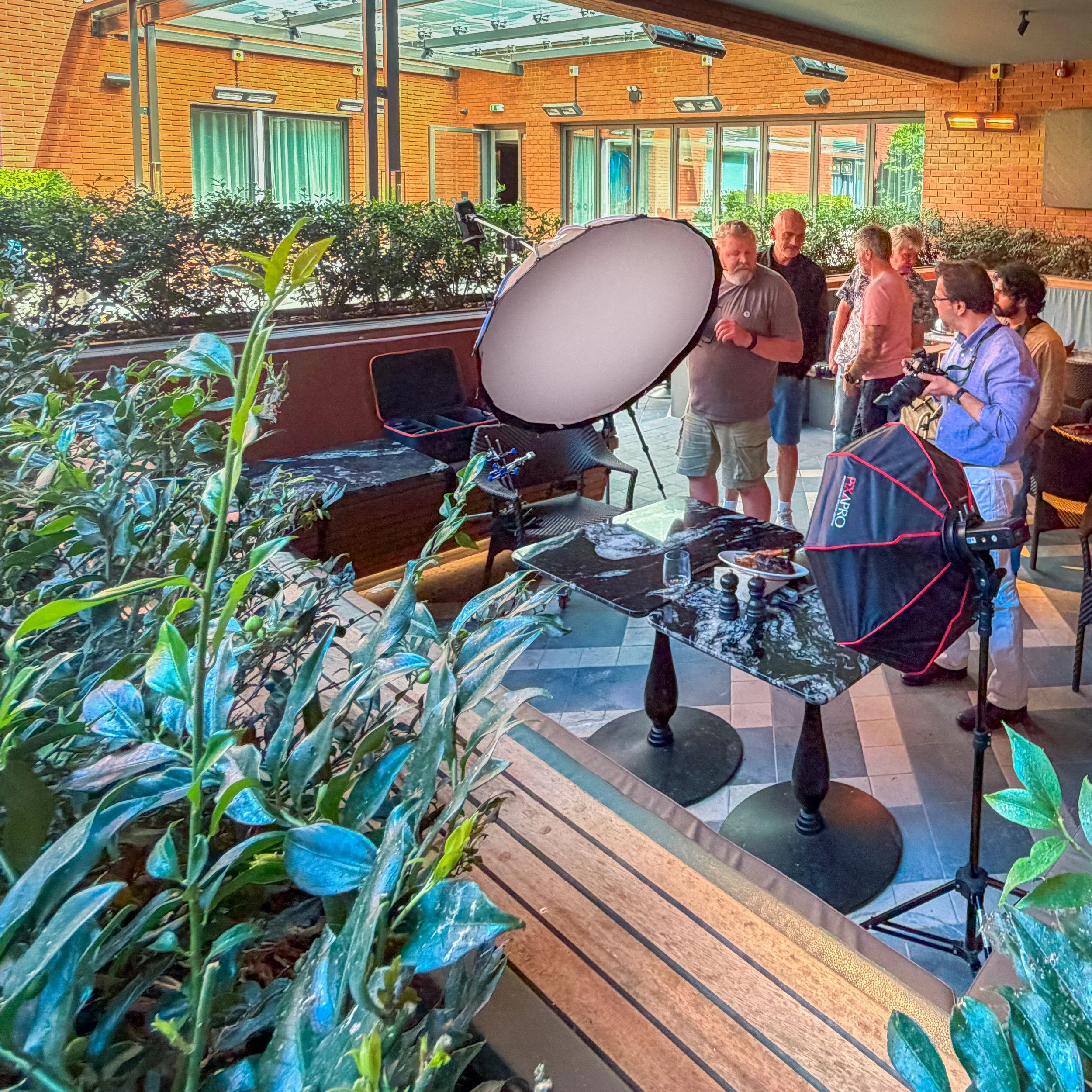 A group of people, mainly older adults, gathered inside a restaurant or café with brick walls, large windows, and greenery outside. There is professional photography equipment such as a large softbox light and camera bags nearby, indicating a photoshoot or filming session.
