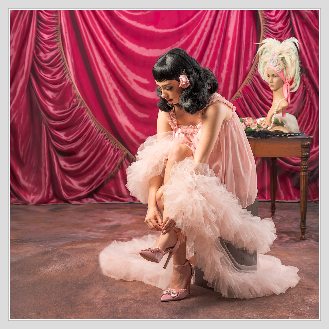A woman in a pink, ruffled dress is sitting on a small platform, adjusting her pink high heels with bows, in front of pink velvet curtains. She has a pink flower in her dark, wavy hair. In the background, a mannequin head is decorated with a large, elaborate feathered headpiece, and a wooden table holds accessories and a bouquet of pink flowers.
