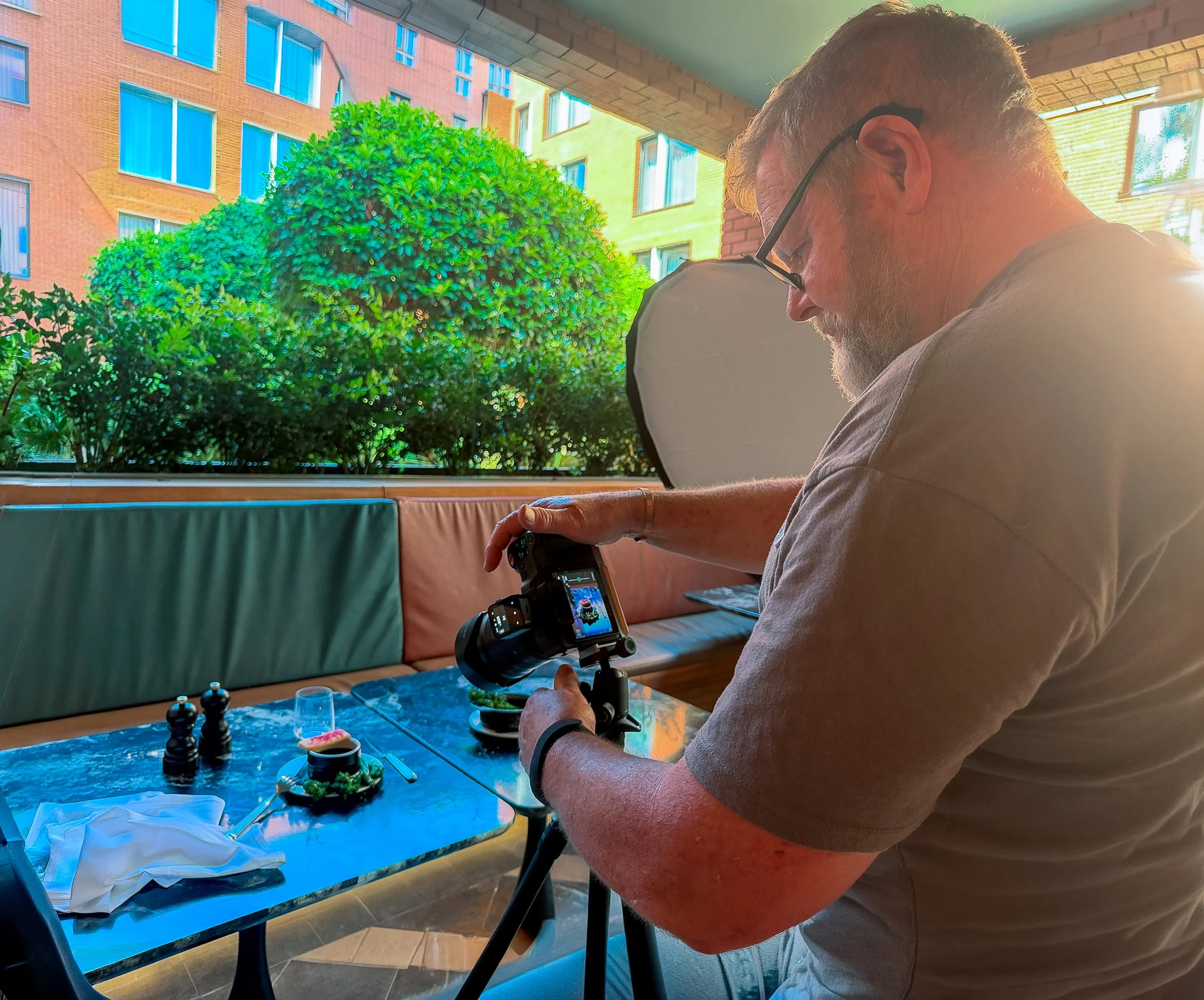 A man with glasses, a beard, and short hair, sitting at a table with a smartphone mounted on a gimbal. The man is looking down at the phone. There are some sushi dishes, soy sauce, and bottles on the table. The background shows large windows with a view of a green tree and multi-story brick and yellow buildings.