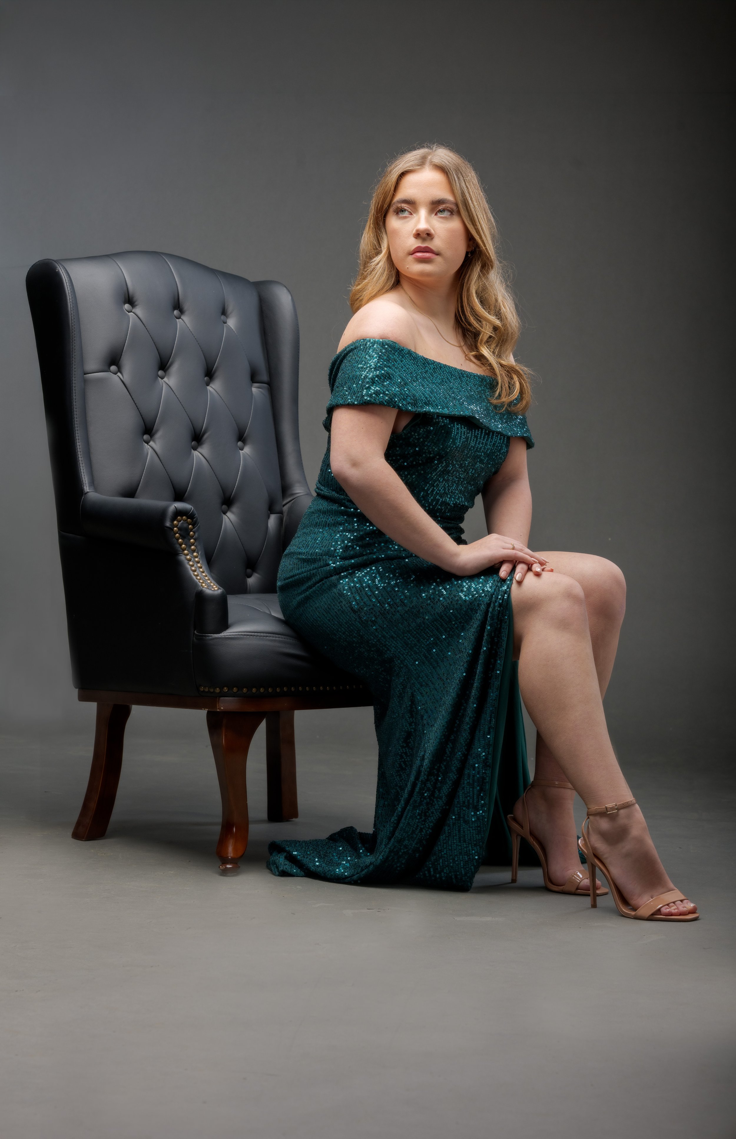 A young woman in a teal, off-the-shoulder, sequined gown sits sideways on the floor, with one knee raised and her hand resting on it. She is wearing high-heeled sandals and is seated next to a dark leather, button-tufted armchair. The background is plain and dark gray.