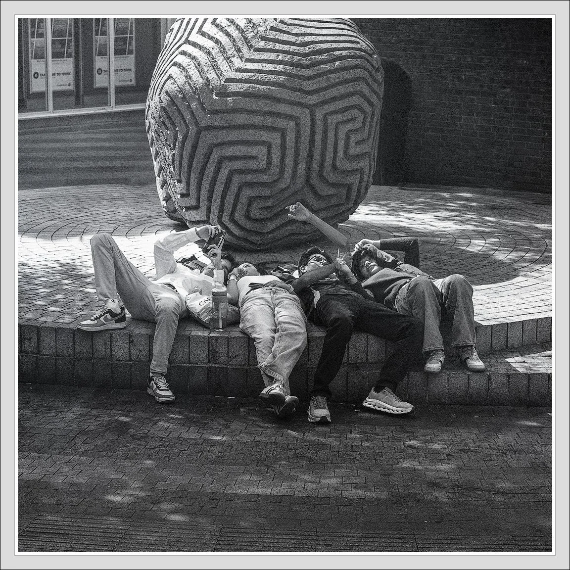 Four young men relaxing and taking selfies in front of a large, maze-patterned sculpture outdoors.