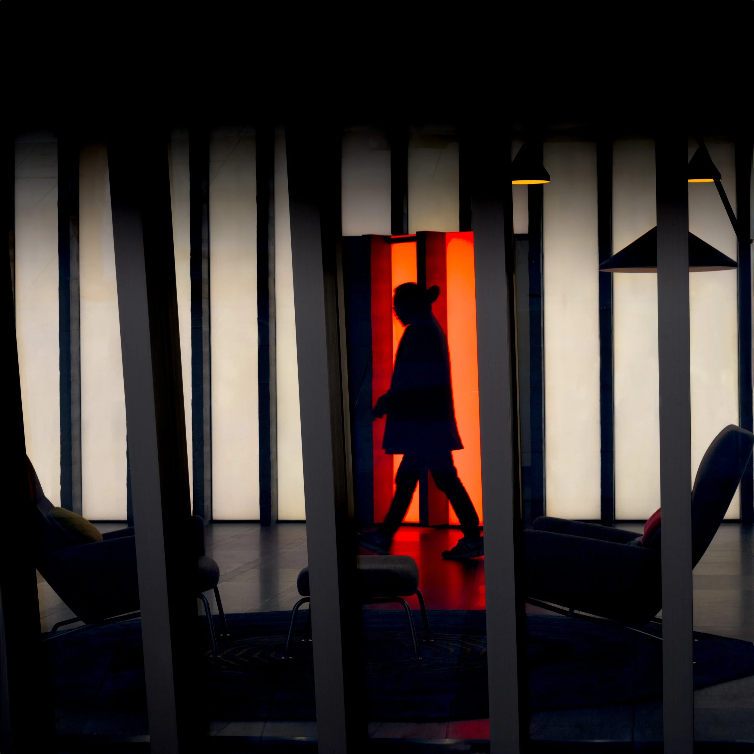 Silhouette of a person walking past a brightly lit vertical red-orange rectangular light fixture in an indoor space with dark vertical panels and hanging lamps.