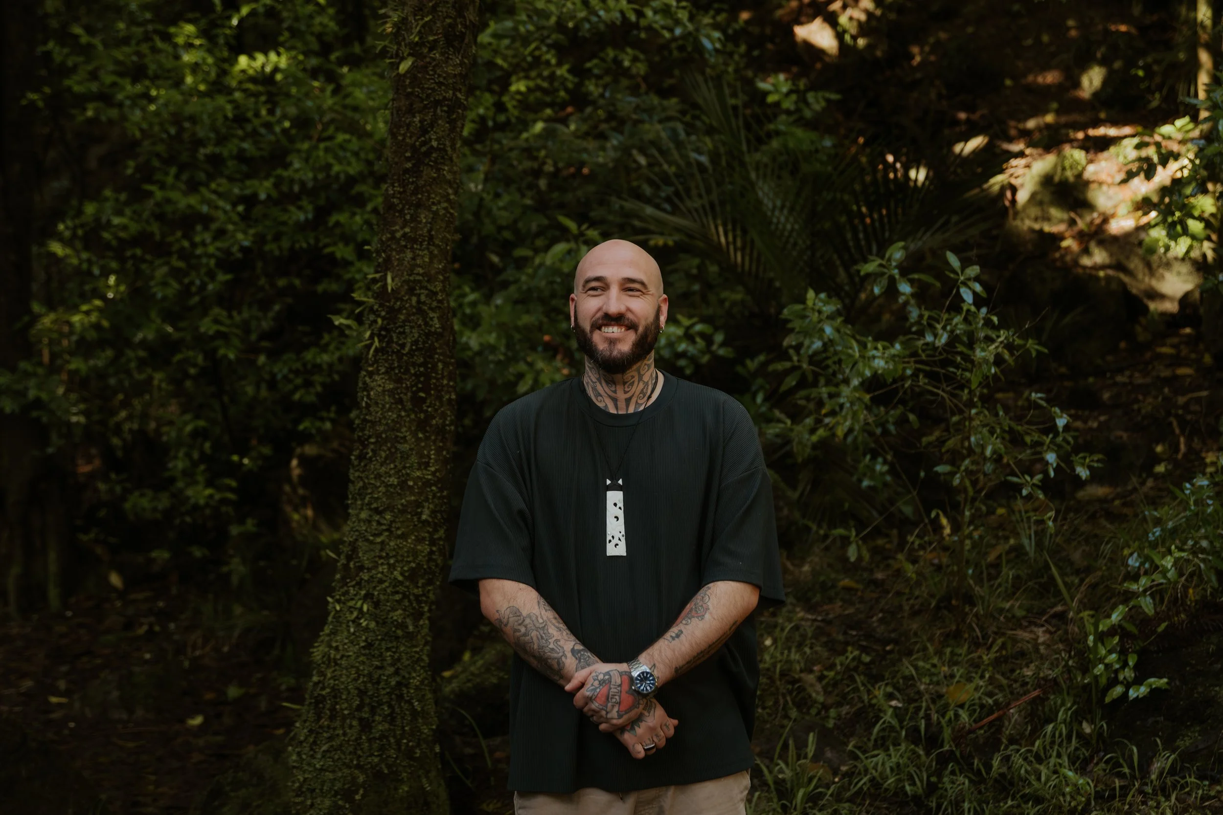 A smiling man with a beard, tattoos on his arms and neck, wearing a black t-shirt, standing outdoors in front of lush green foliage and trees.