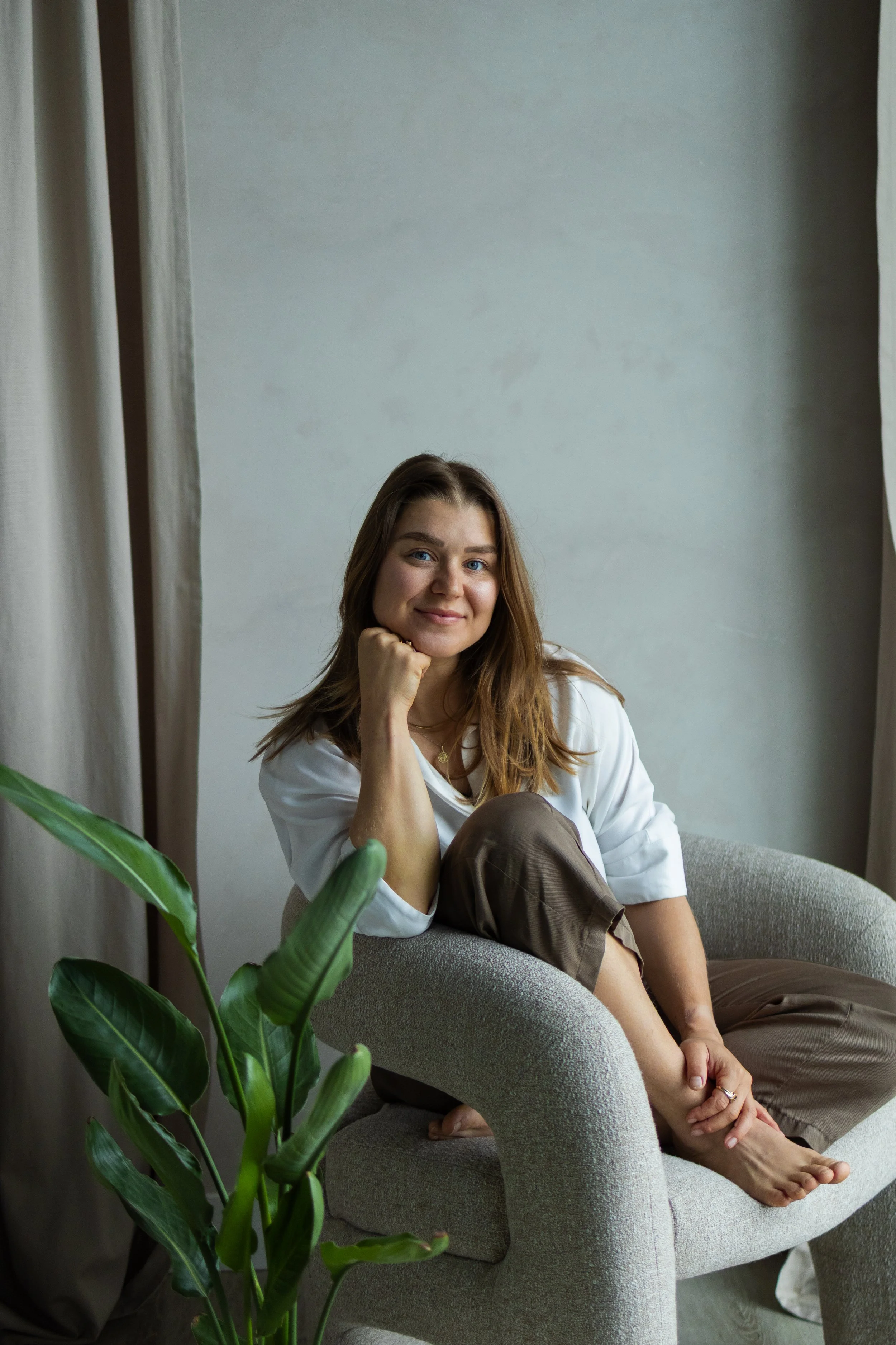 A young woman with long hair sitting casually on a gray armchair in a living room, smiling at the camera, with a large green plant in the foreground and beige curtains behind her.