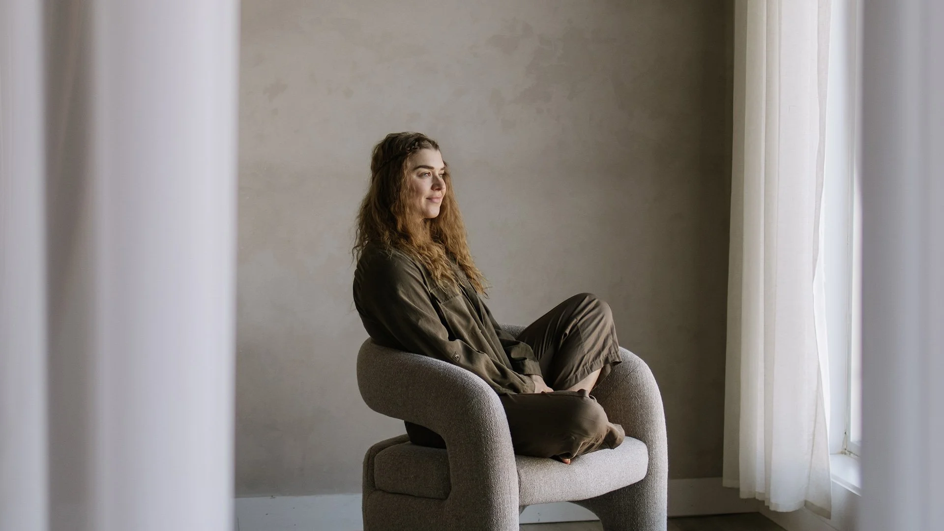 A woman with long curly hair sitting on a gray armchair, looking out a window with white curtains in a minimalist room with plain beige walls.