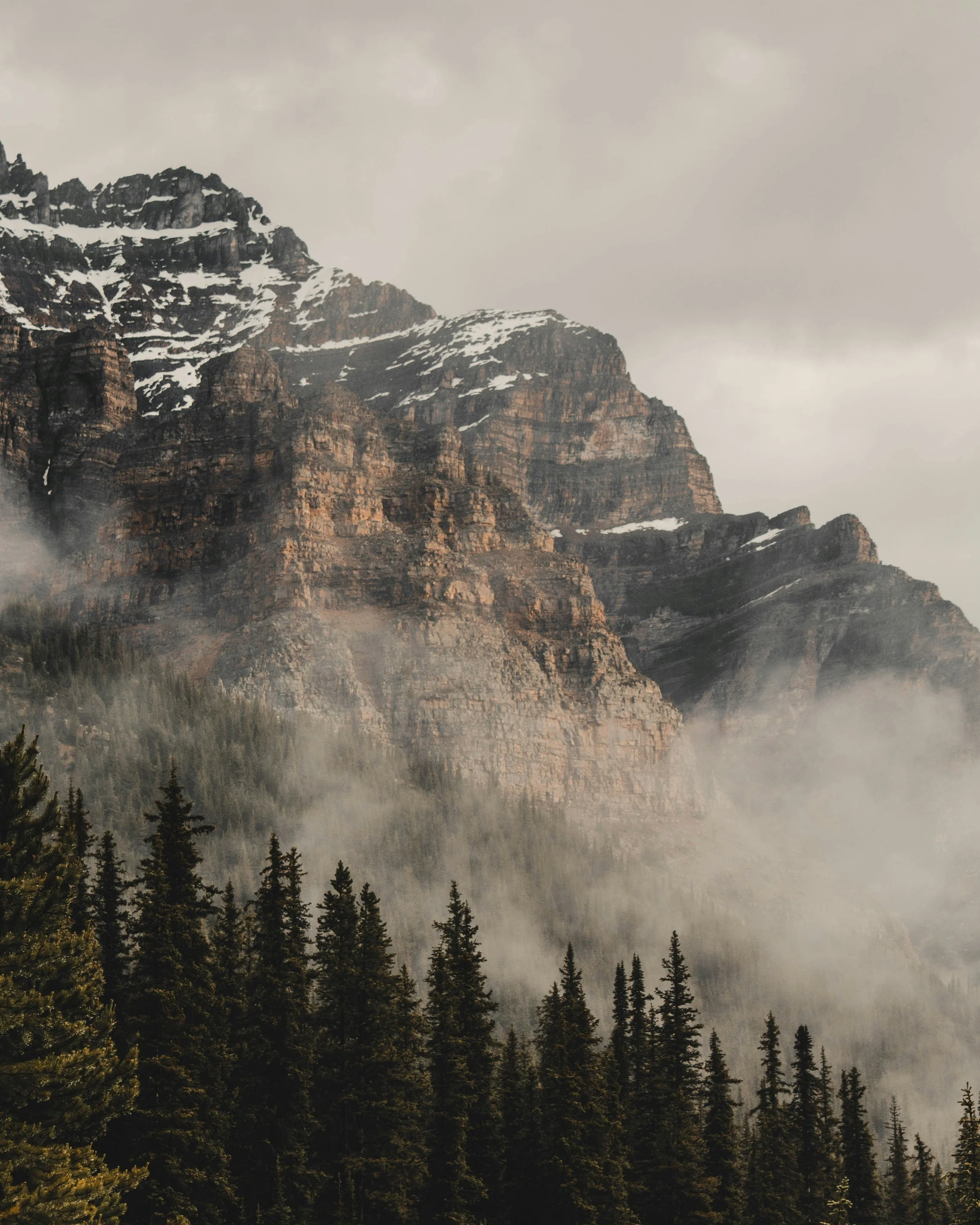 A rugged mountain with patches of snow on its peaks, surrounded by a dense evergreen forest, with fog at the base and cloudy sky overhead.