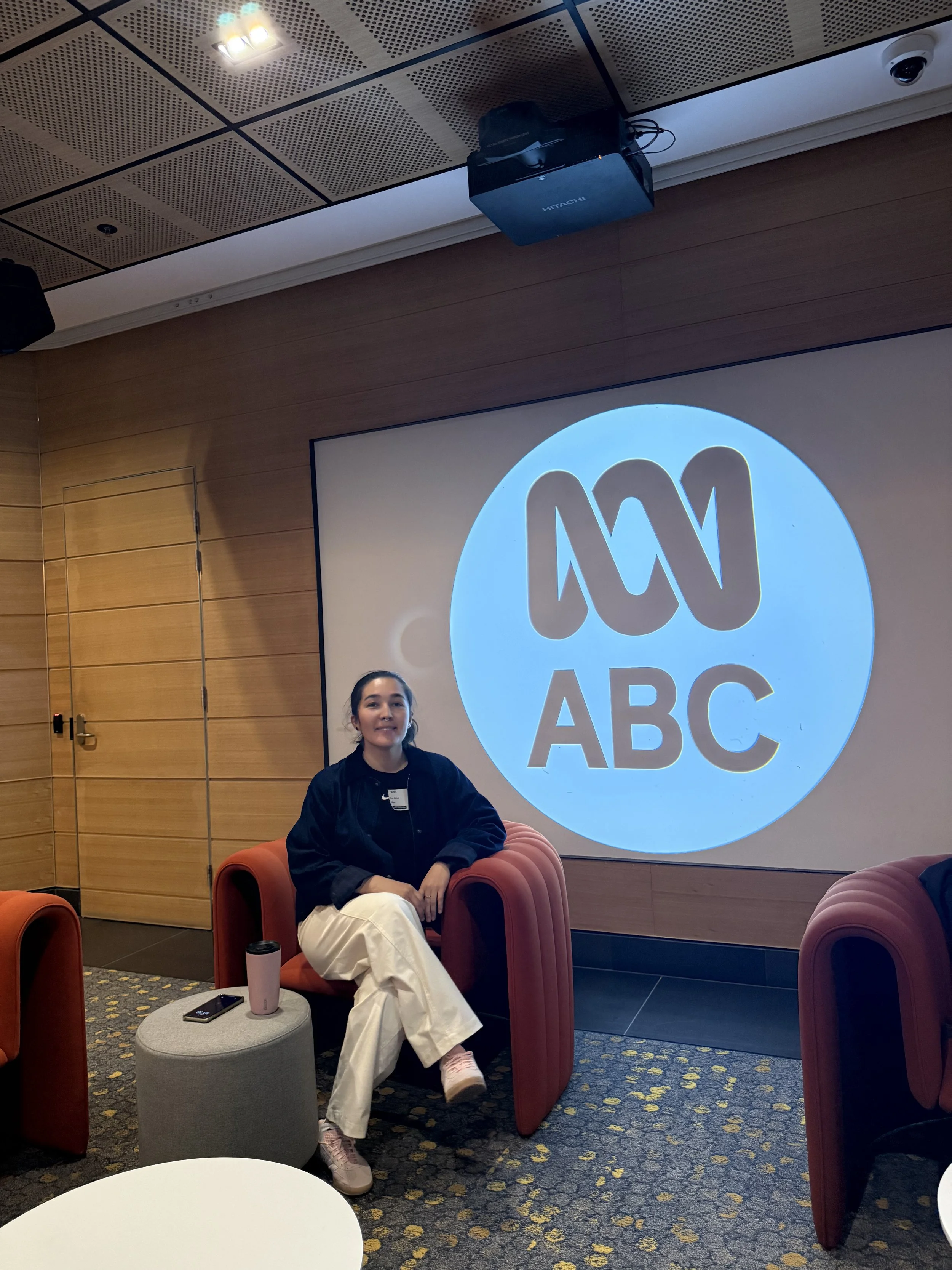A young woman sitting on a red armchair in a modern conference room with wooden panel walls. Behind her, a large screen displays the ABC logo, and there are several orange chairs and a small table with a pink tumbler, a black phone, and a white cylin